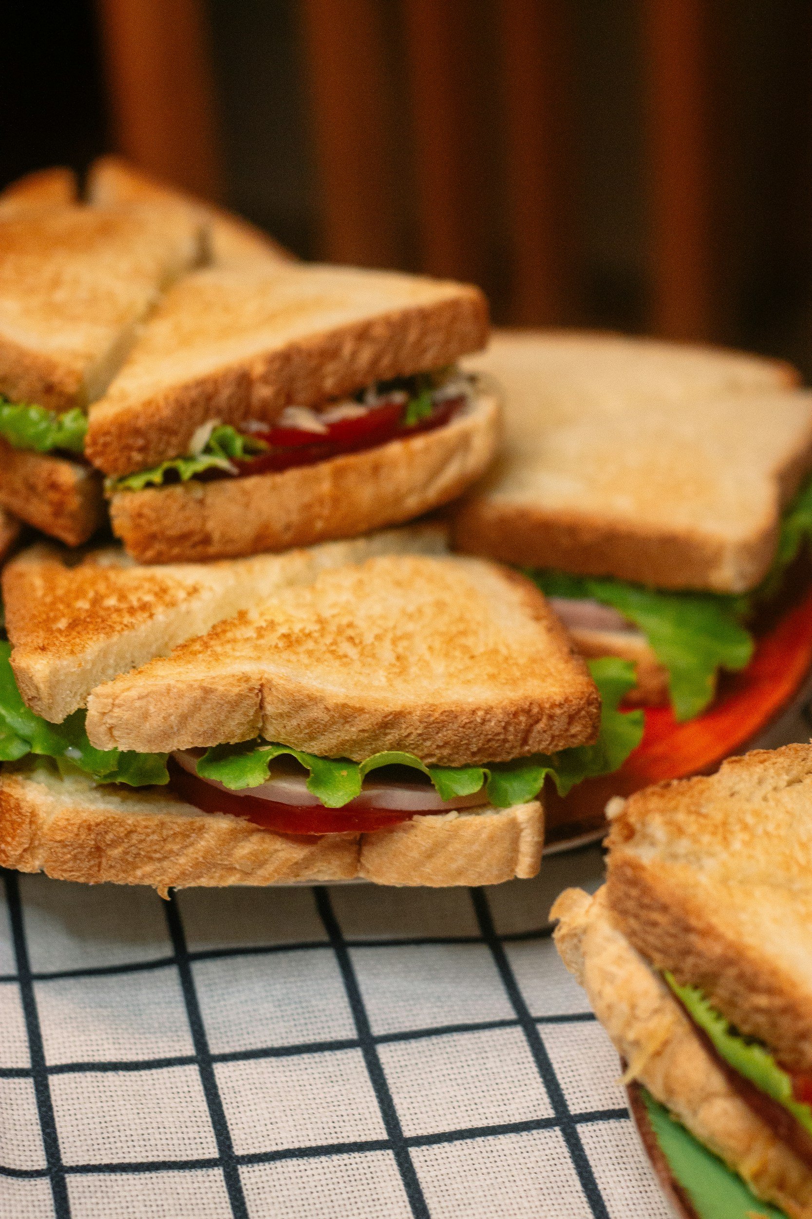 Close-up of toasted sandwiches with lettuce, tomato, and possibly turkey or chicken, on a wire rack with a black and white checkered cloth underneath.
