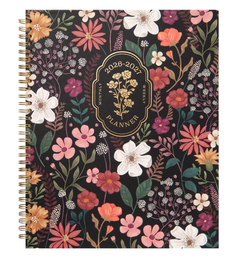 A floral-patterned planner with a black background, decorated with various pink, white, peach, and purple flowers and green leaves. The cover features a golden emblem that reads "2026-2027 Monthly & Weekly Planner".
