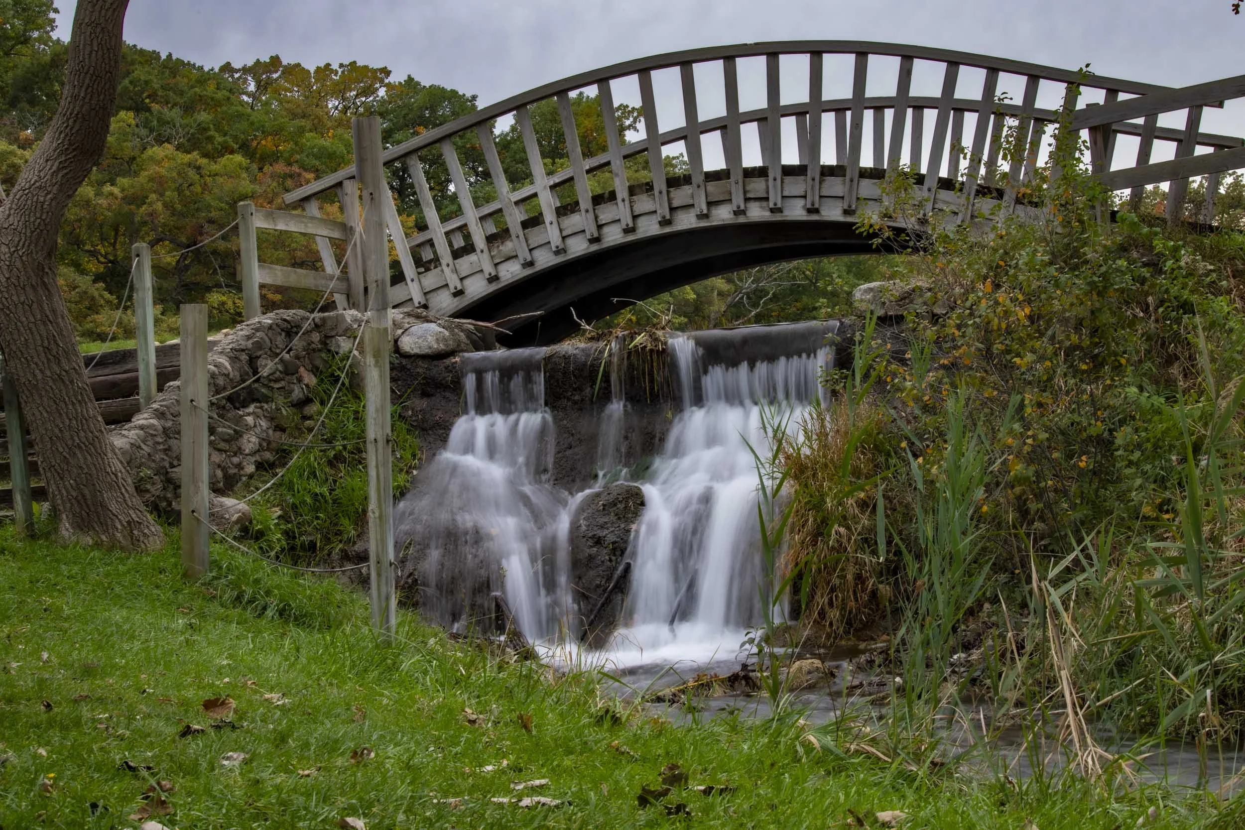 A small waterfall flowing beneath an arched wooden footbridge beside a grassy bank.