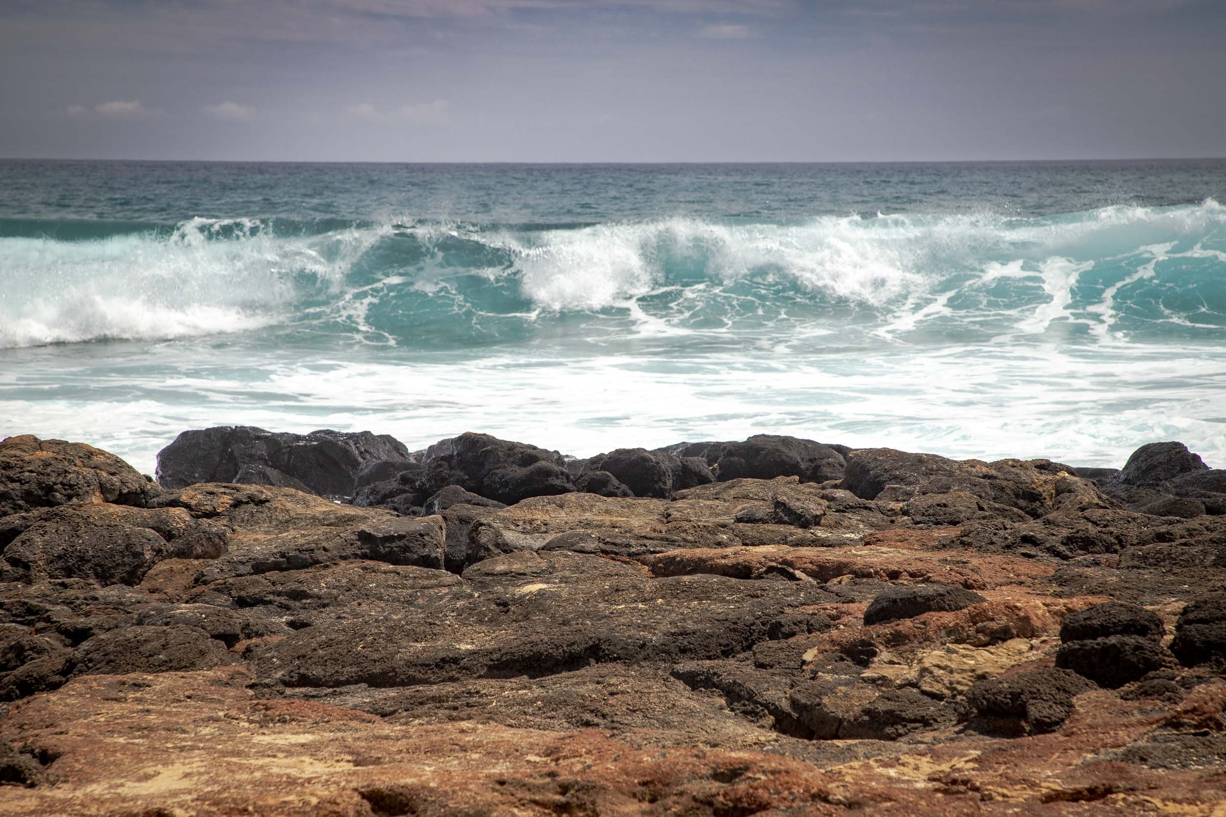 Turquoise waves breaking beyond rugged dark coastal rocks.