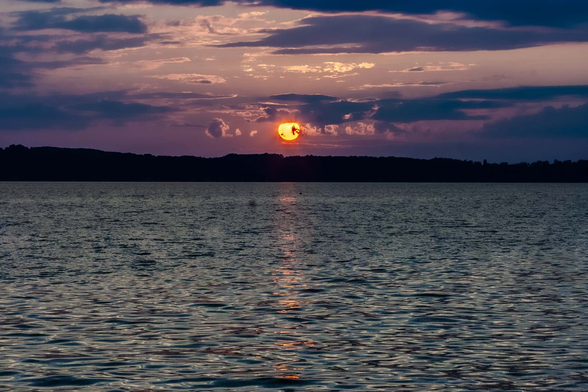 Sun breaking through purple and blue clouds above a lake at dusk.