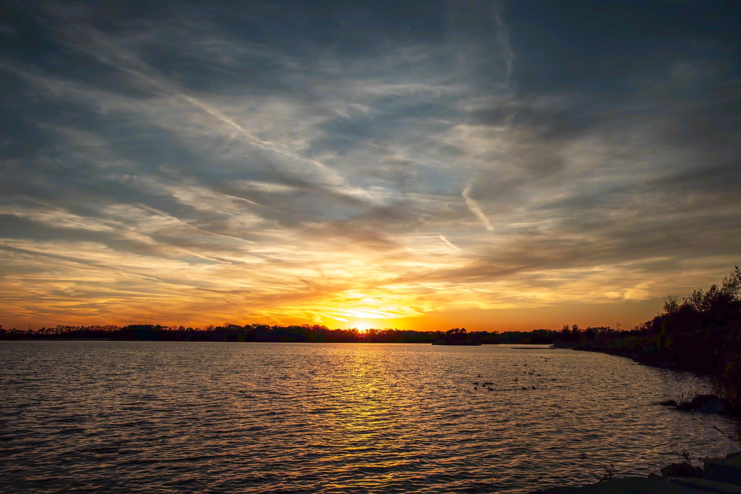 Golden sun setting over rippled lake water beneath streaks of gray and orange clouds.