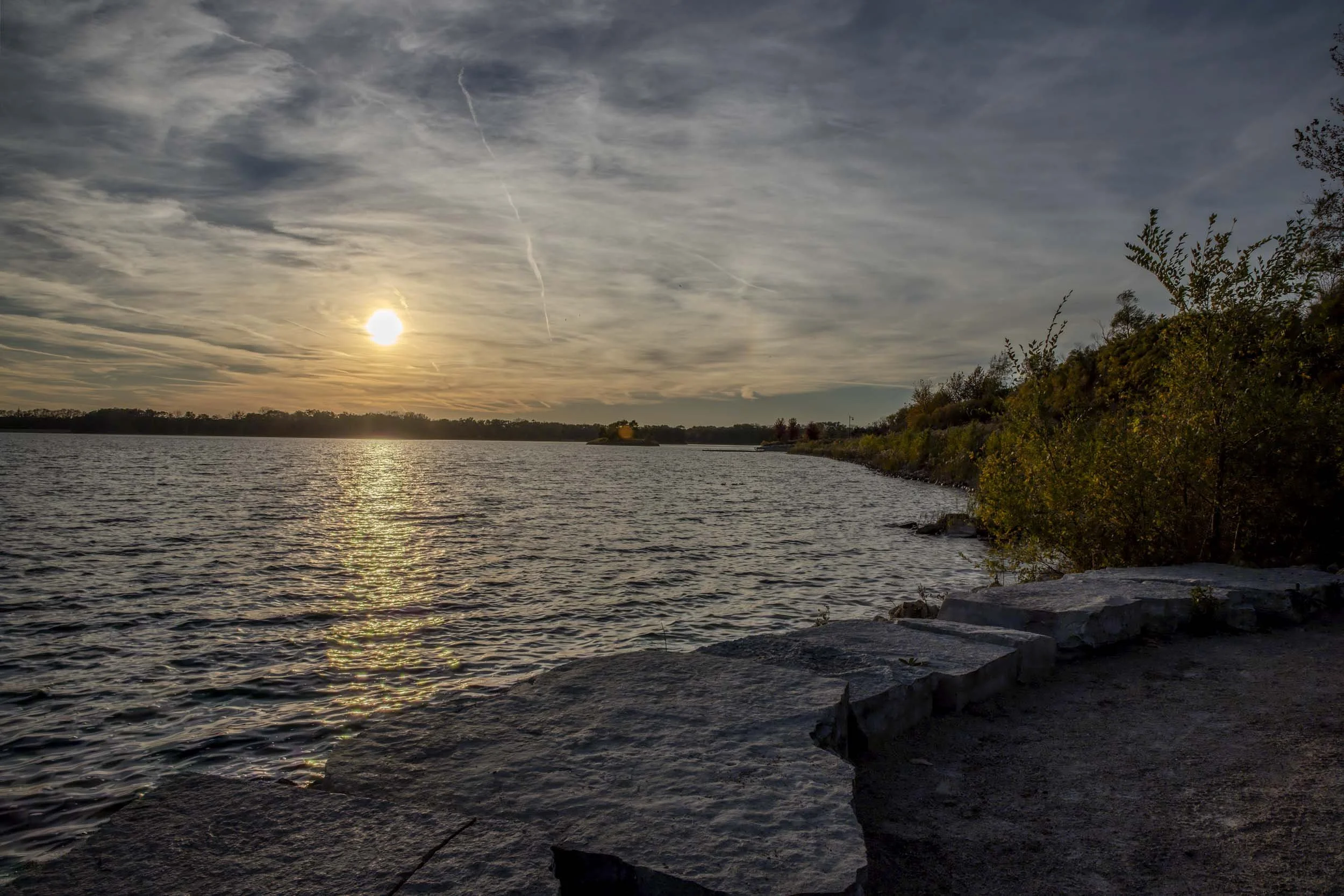 Low sun reflecting across a lake beside a rocky shoreline under a cloudy sky.
