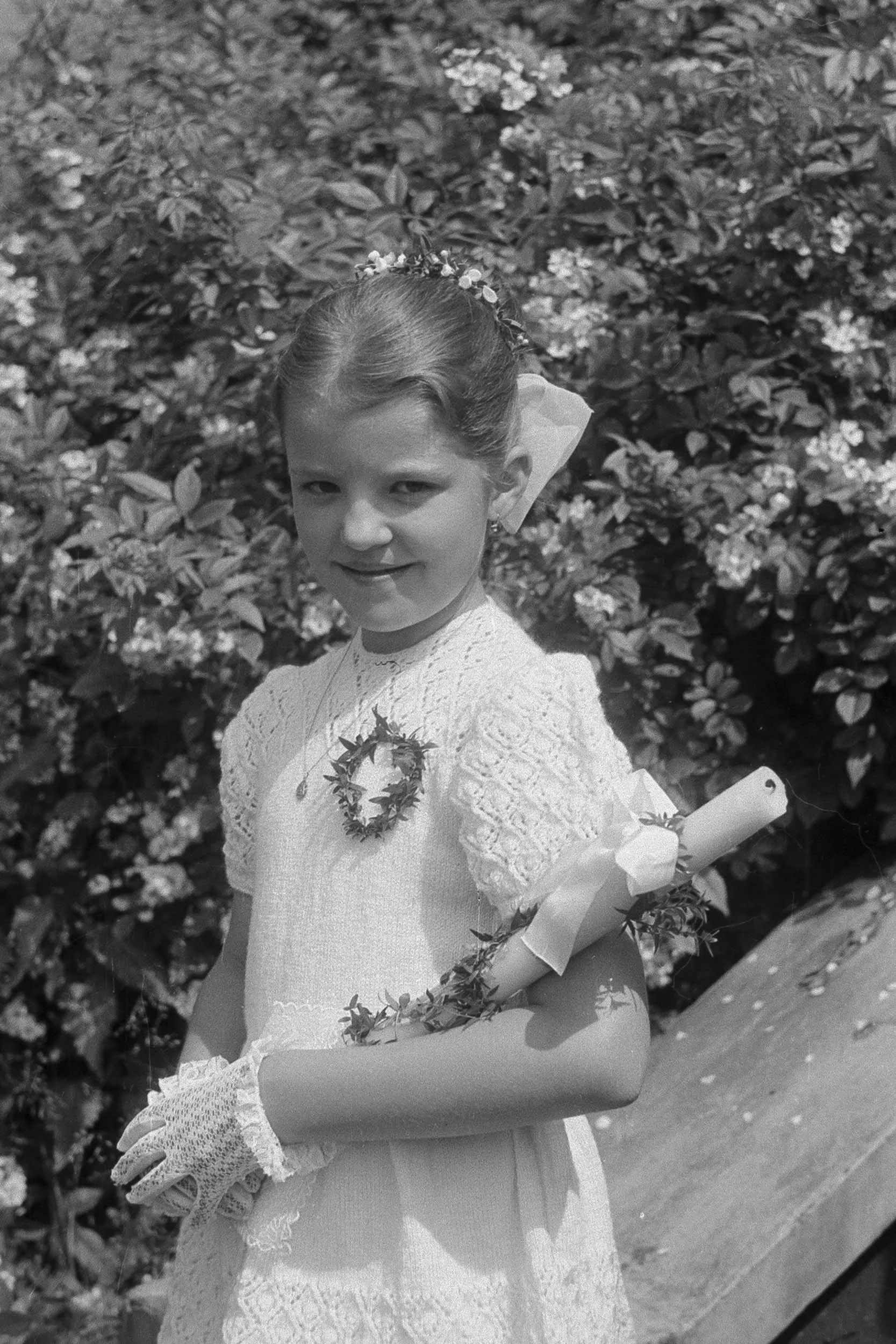 Restored black-and-white photograph of a girl wearing a dress and gloves, holding a rolled document beside flowering shrubs.