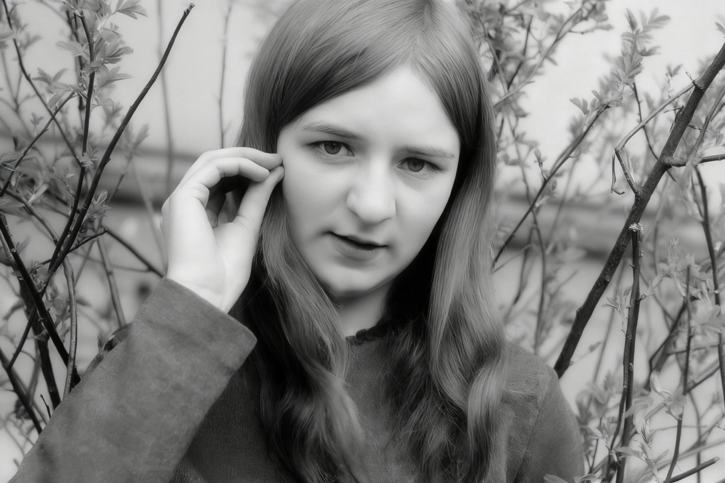 Black-and-white portrait of a young woman touching her hair beside bare branches.
