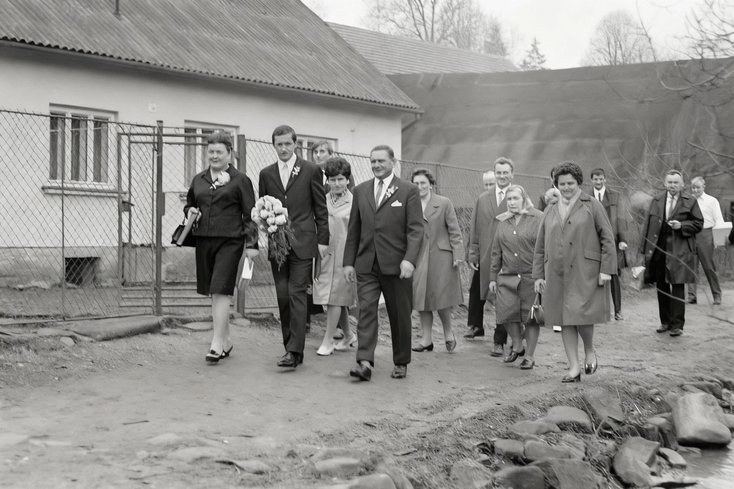Black-and-white photograph of a group in formal clothing walking along an unpaved road beside houses.
