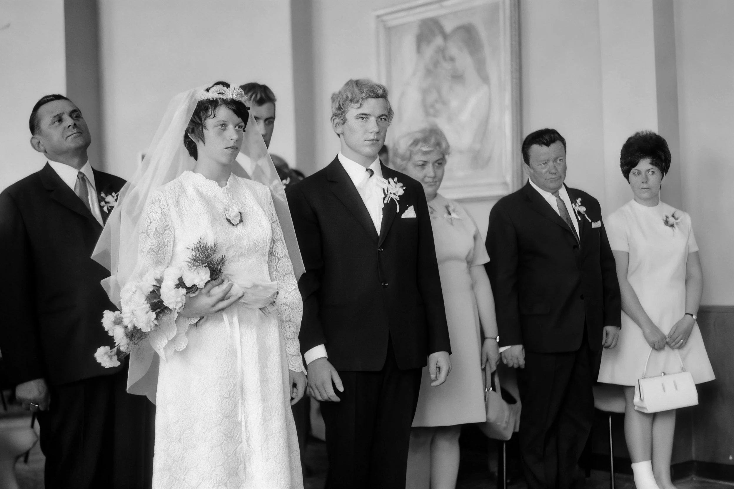 Black-and-white photograph of a bride holding a bouquet beside a groom and wedding guests indoors.