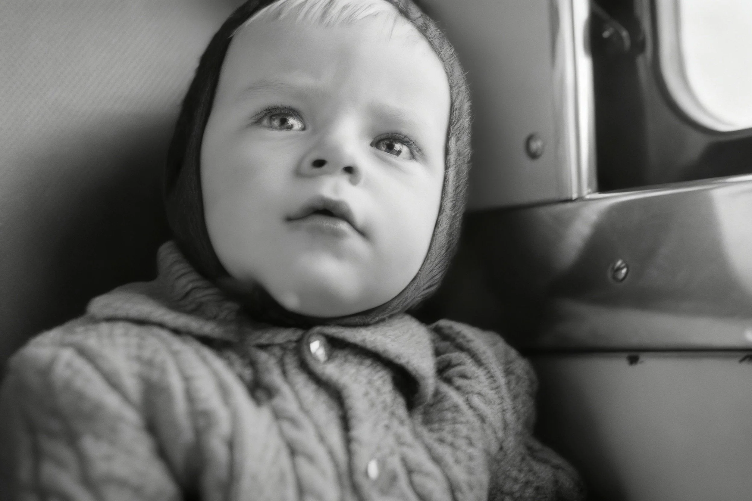 Black-and-white photograph of a small child in a knitted hooded sweater looking upward inside a vehicle.