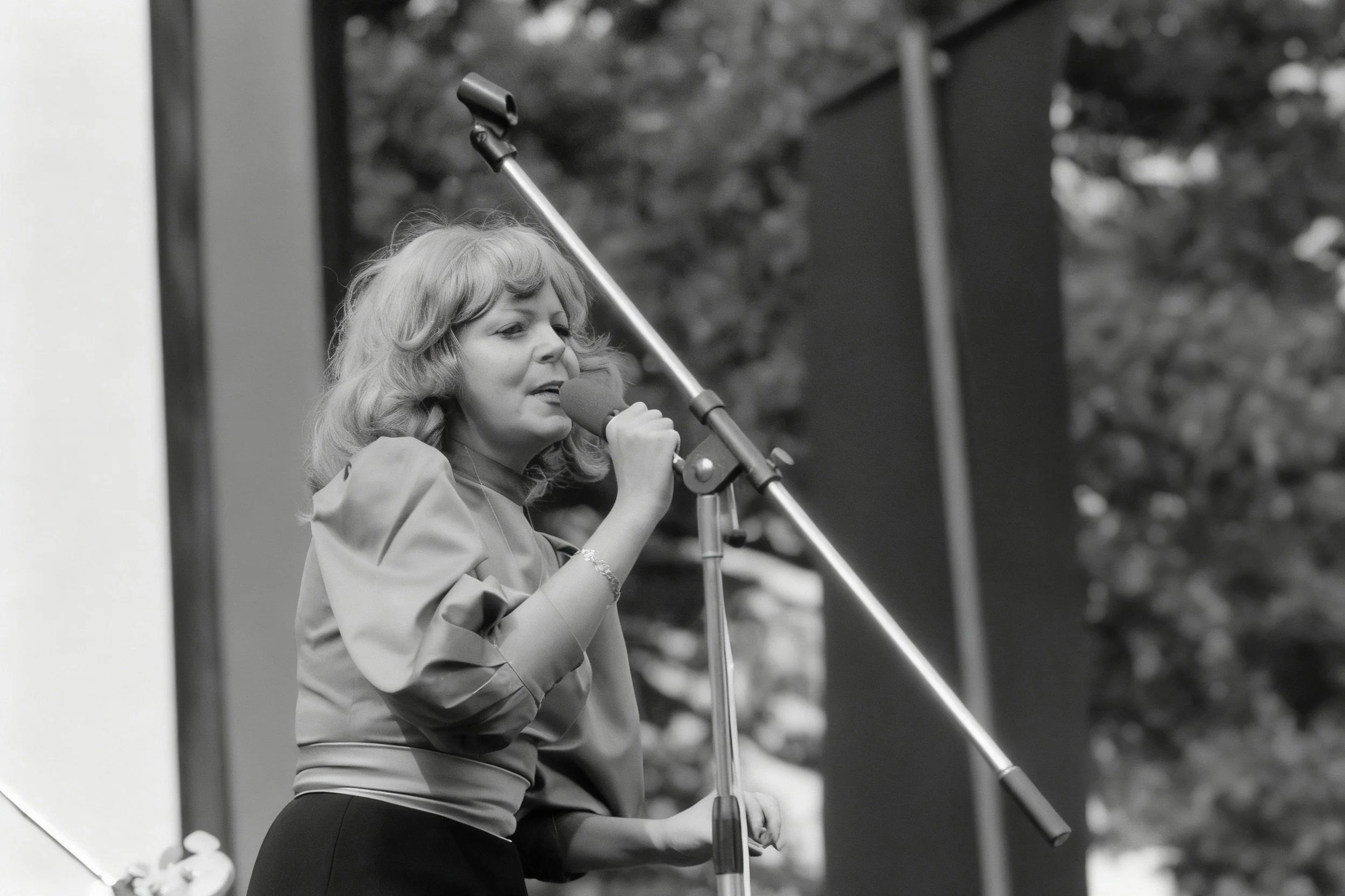 Black-and-white photograph of a woman singing into a handheld microphone on an outdoor stage.