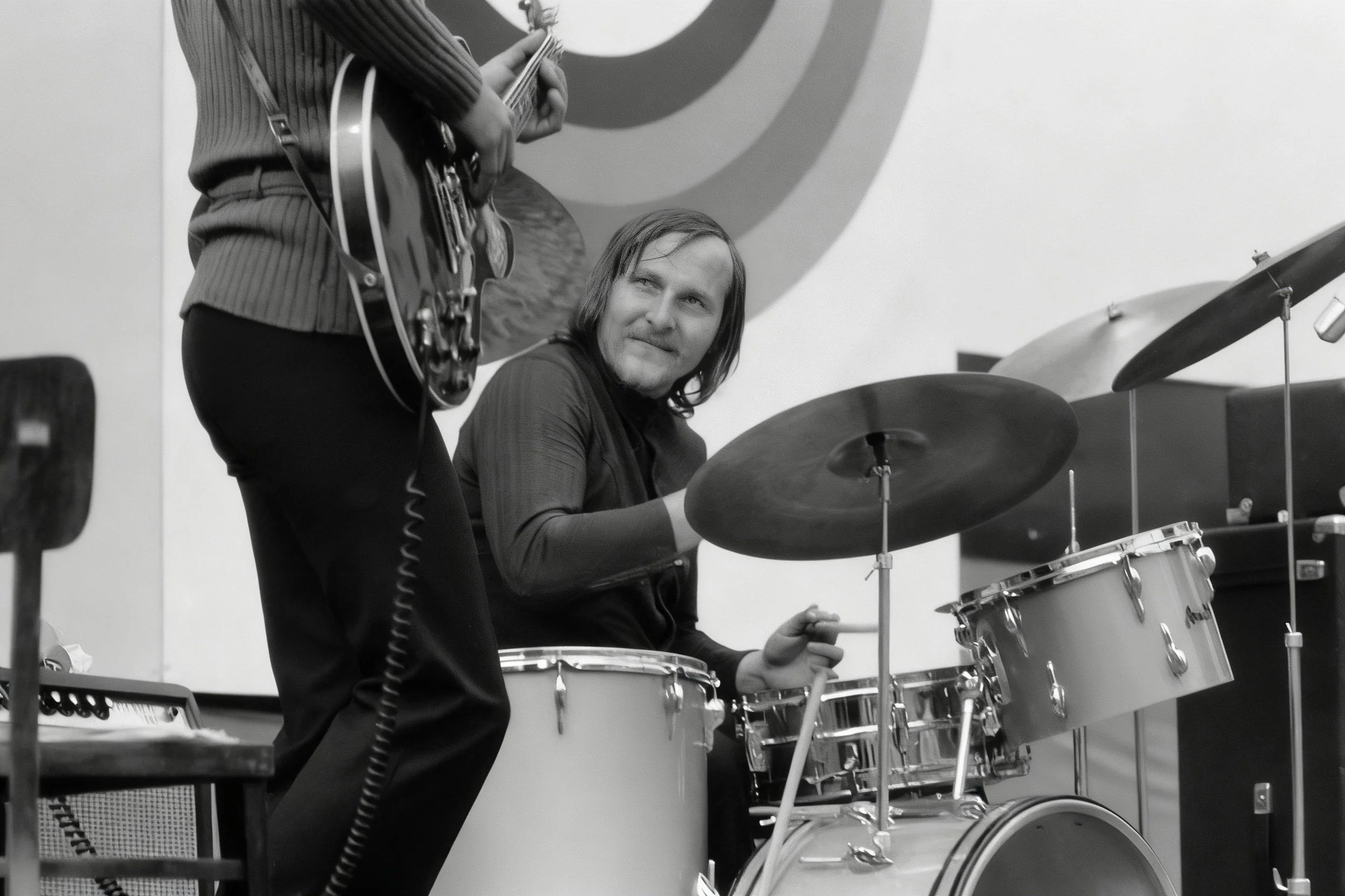 Black-and-white photograph of a drummer behind a drum kit, with a guitarist partly visible in the foreground.