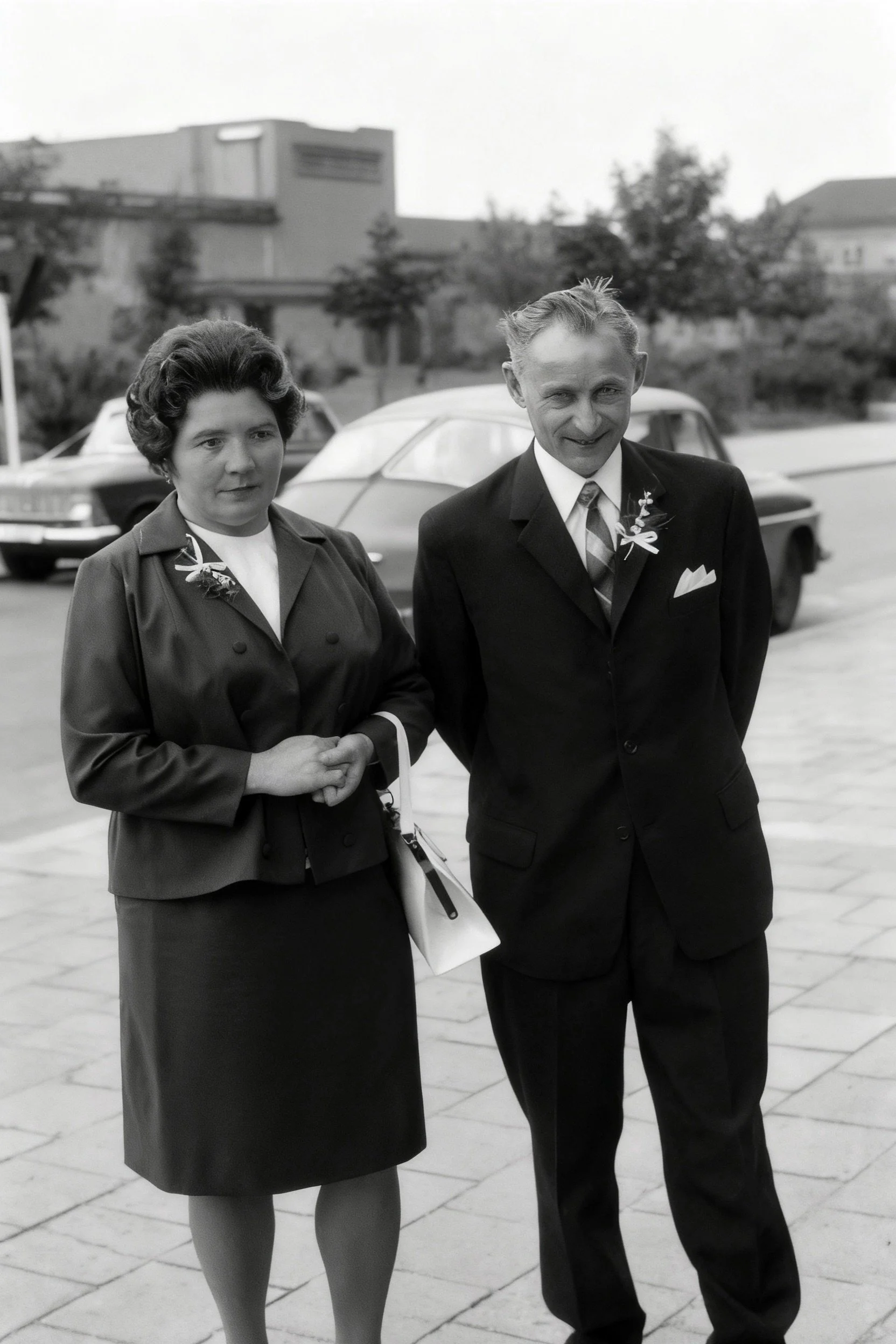 Black-and-white photograph of a woman and a man in formal clothing standing arm in arm on a paved square.