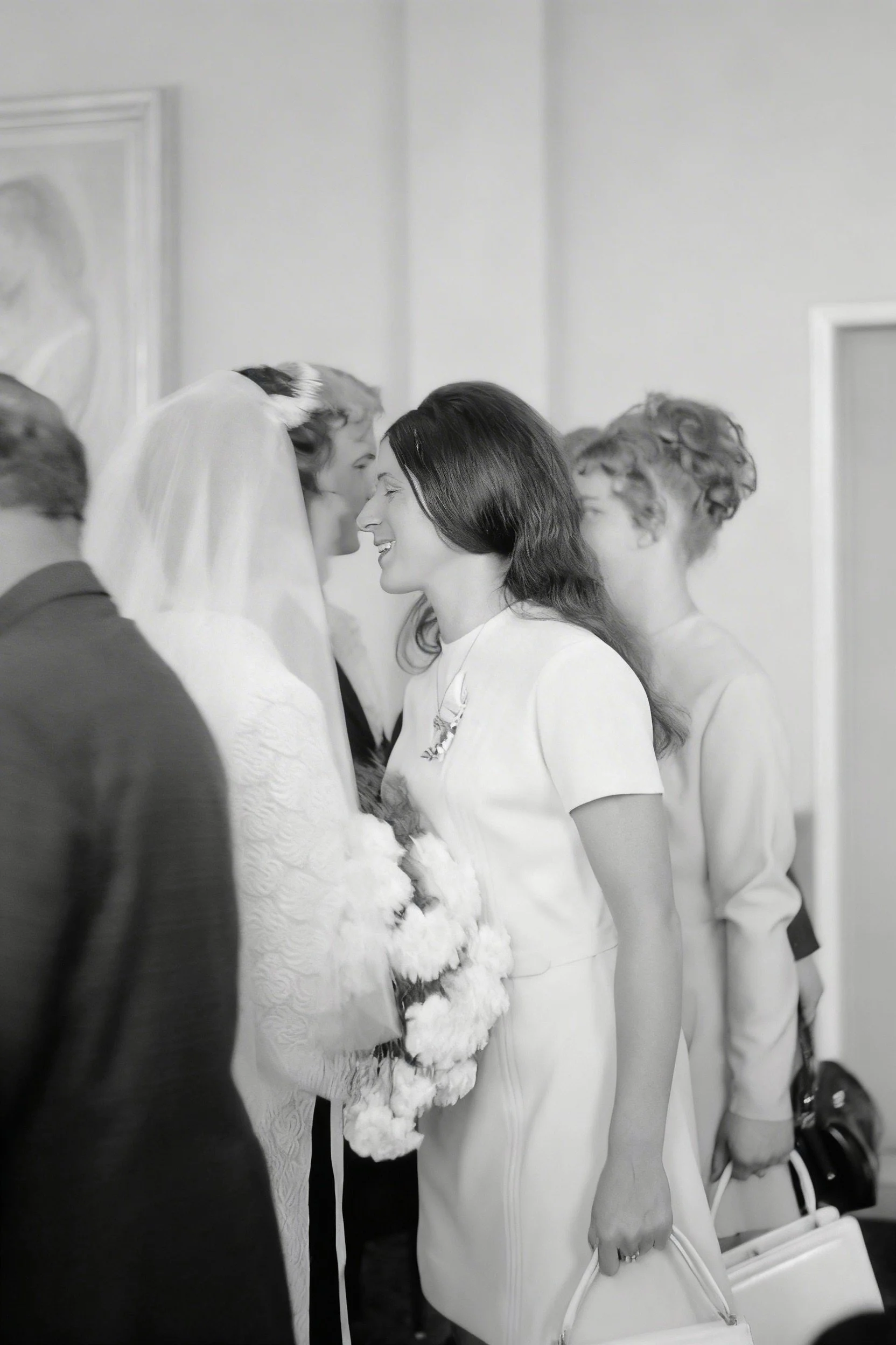 Black-and-white photograph of a bride in a veil greeting a woman among guests indoors.