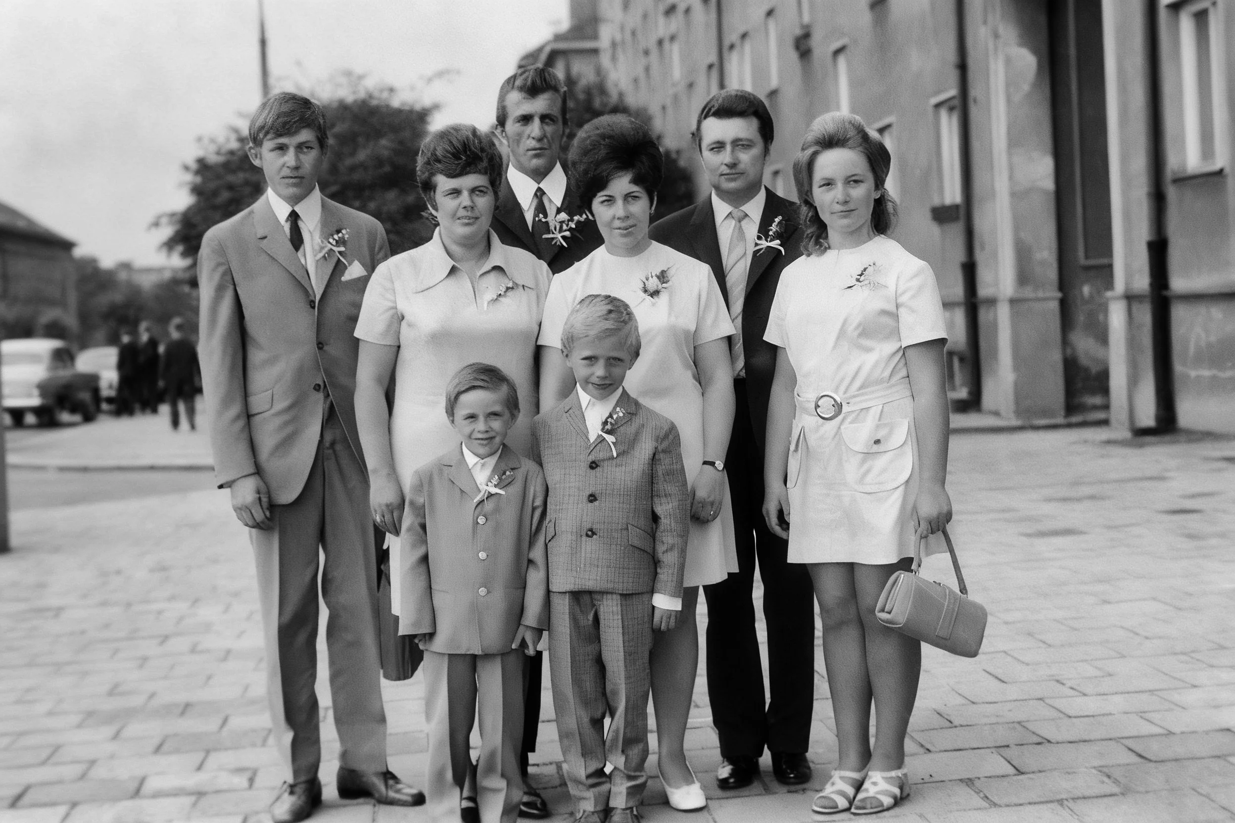 Black-and-white group portrait of adults and two boys in formal clothing on a paved square.
