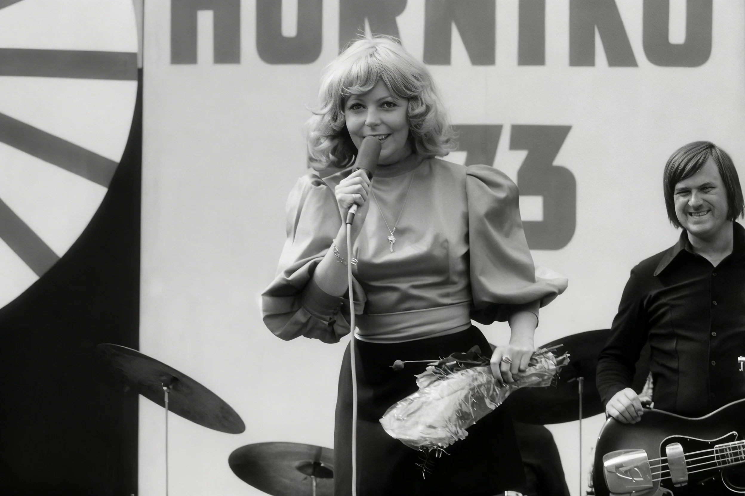 Black-and-white photograph of a woman singing into a microphone beside a bass guitarist onstage.