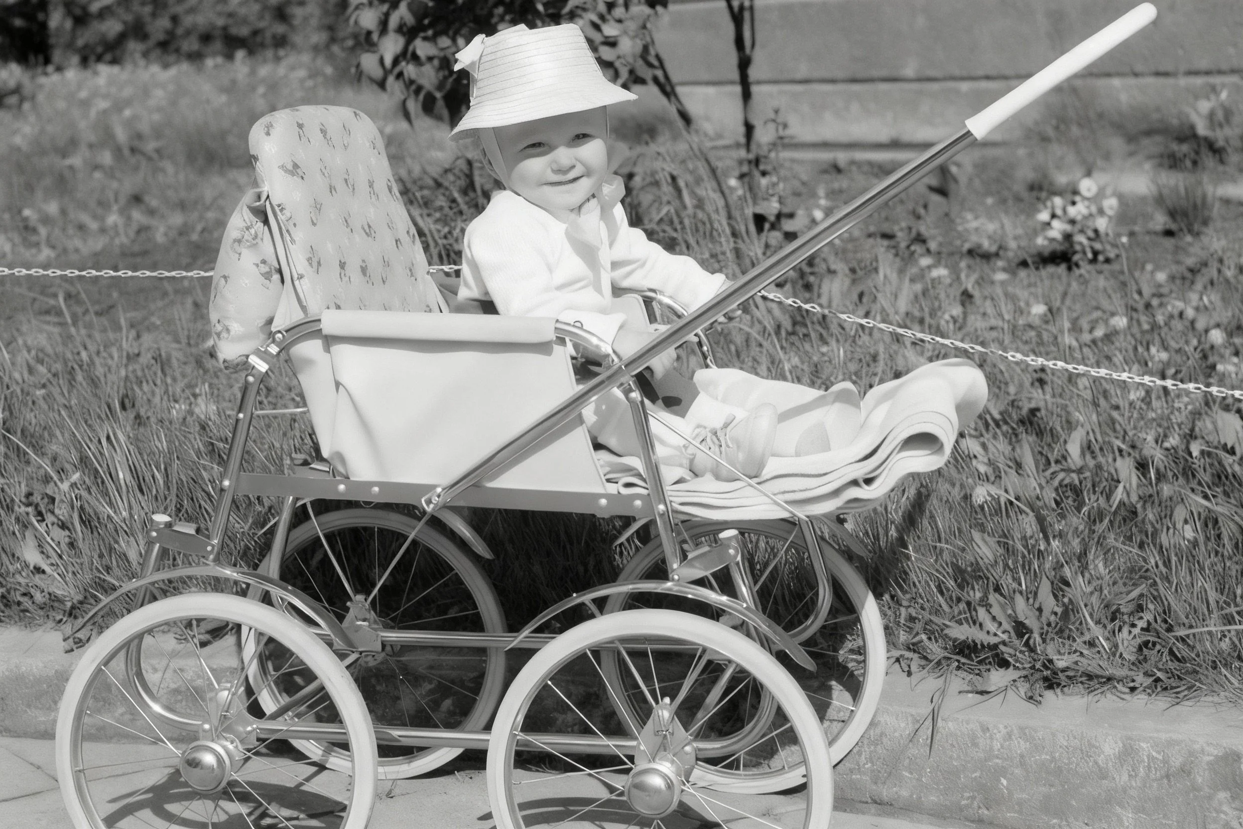 Black-and-white photograph of a small child wearing a light-colored hat seated in a pram outdoors.
