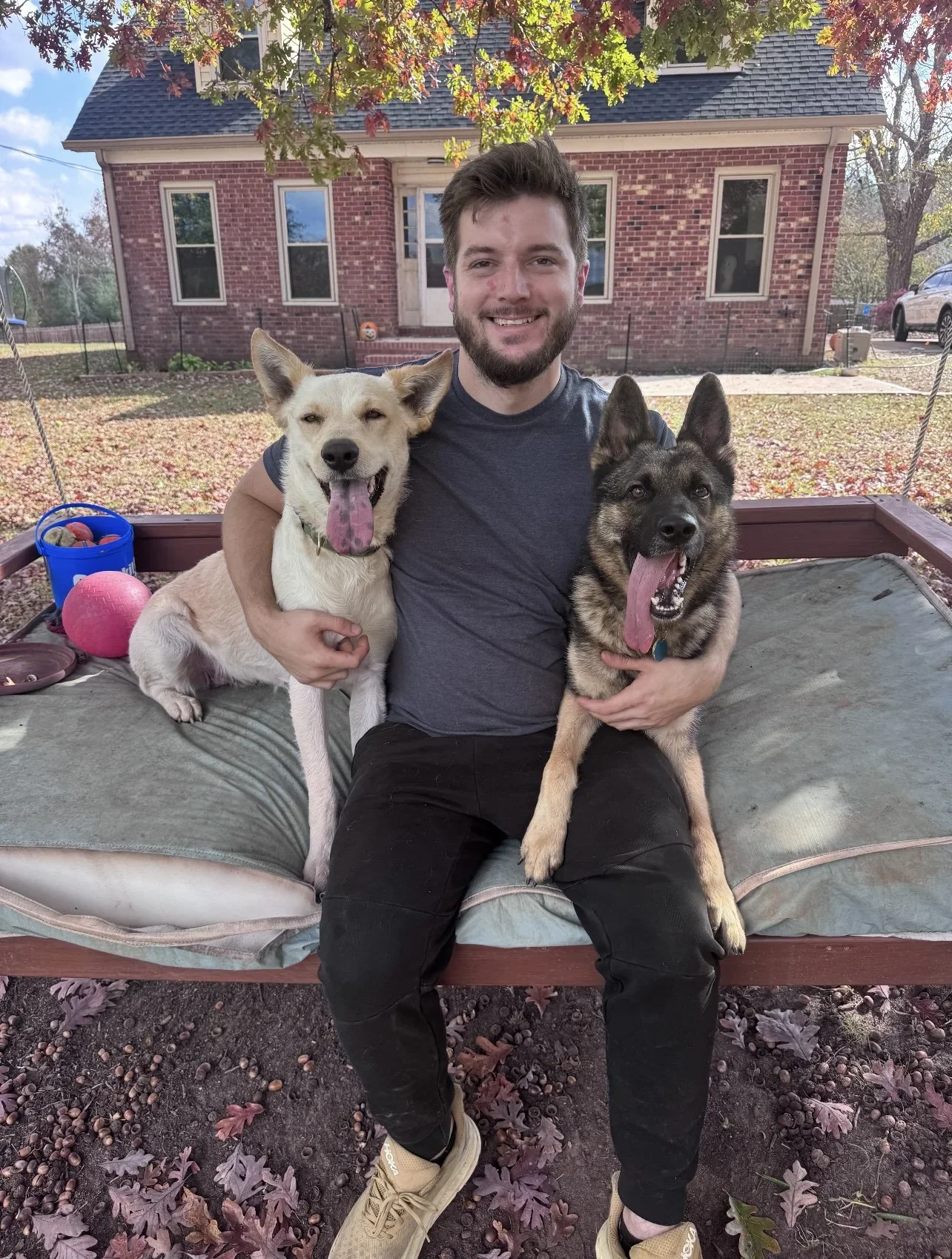 A man sitting on a bench with two dogs, one white and one black and tan, outdoors in front of a brick house with autumn trees and fallen leaves.
