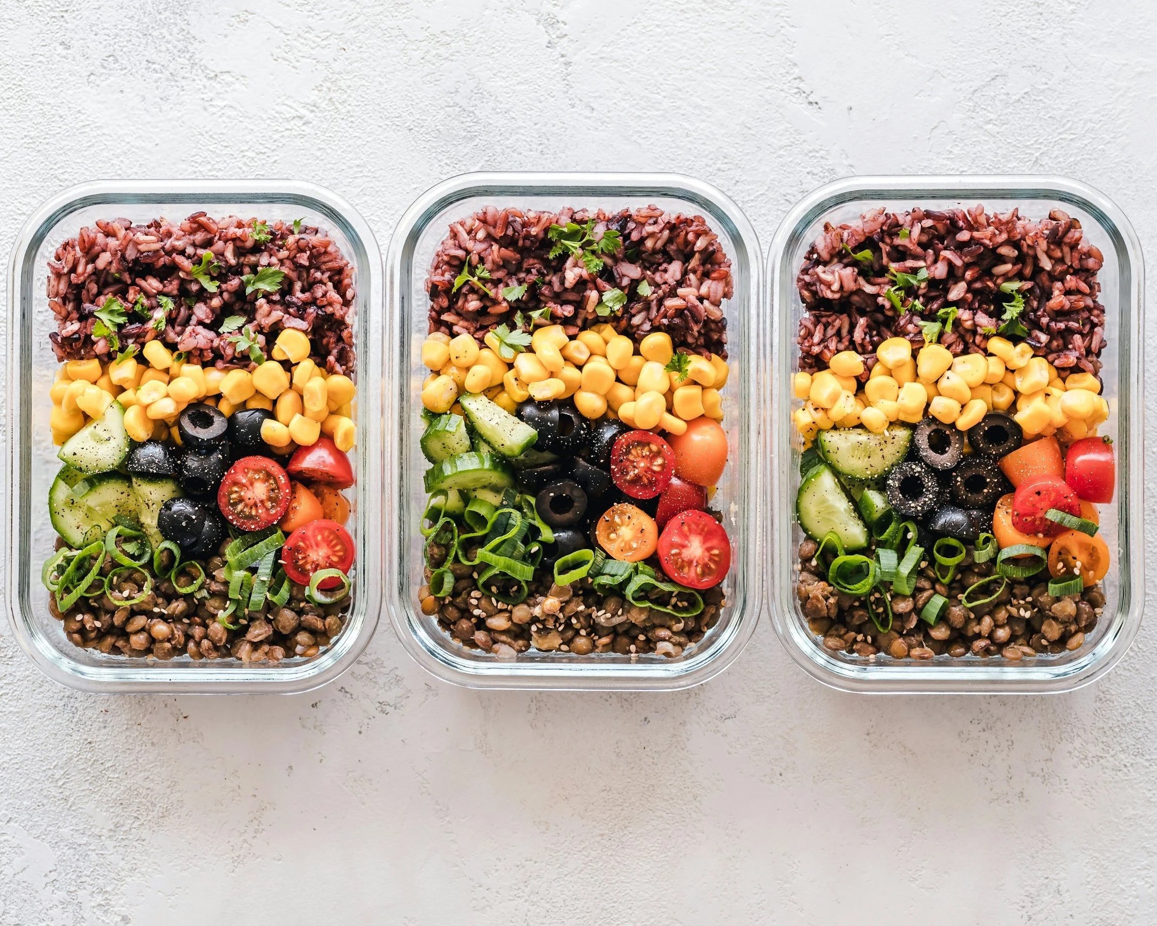 Three glass meal prep containers filled with layered salad ingredients including cherry tomatoes, cucumber slices, black olives, chickpeas, cooked lentils, and garnished with parsley and green onions, placed on a white textured surface.