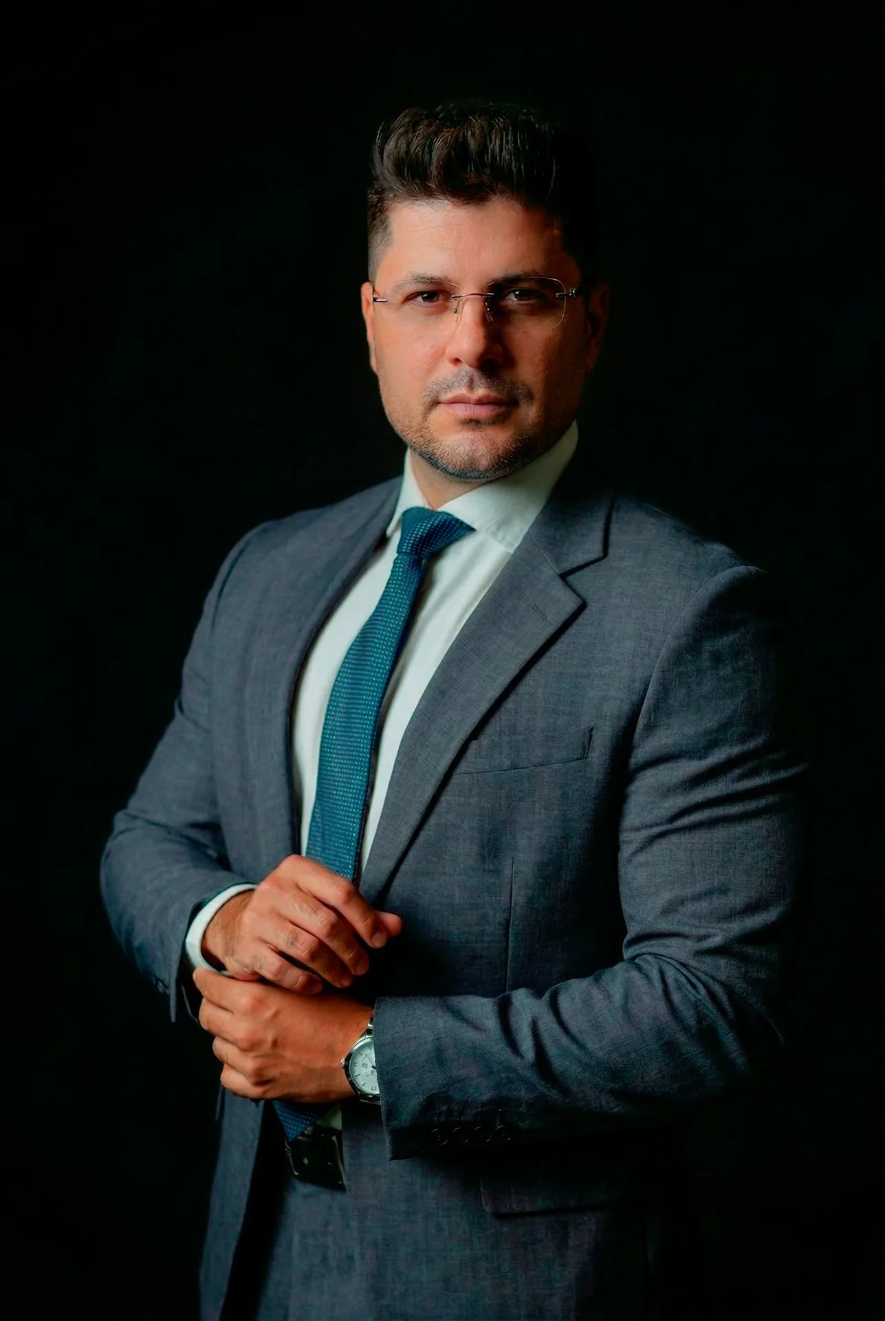 Homem de terno cinza, camisa branca e gravata azul escura, usando óculos e relógio, posando com fundo preto.