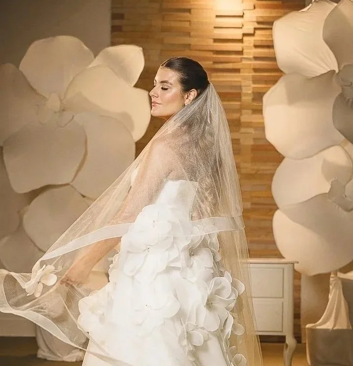 Noiva em vestido branco com detalhes de flores, usando véu, em cenário decorado com grandes flores brancas e parede de madeira ao fundo.