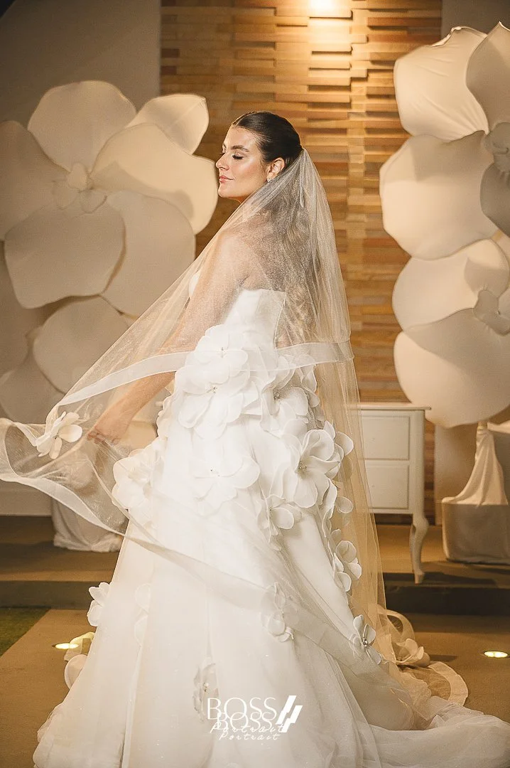 Noiva em vestido branco com detalhes de flores, usando véu, em cenário decorado com grandes flores brancas e parede de madeira ao fundo.