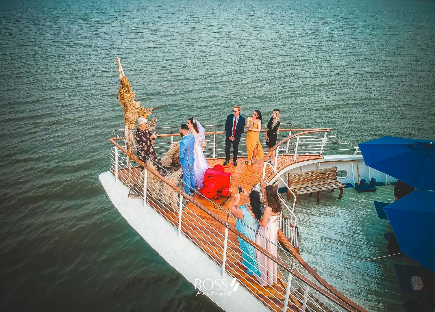 Casal de noivos em cerimônia de casamento na suíte de um barco, com convidados ao redor na parte superior ao ar livre, painel de fundo decorado, e o mar ao redor