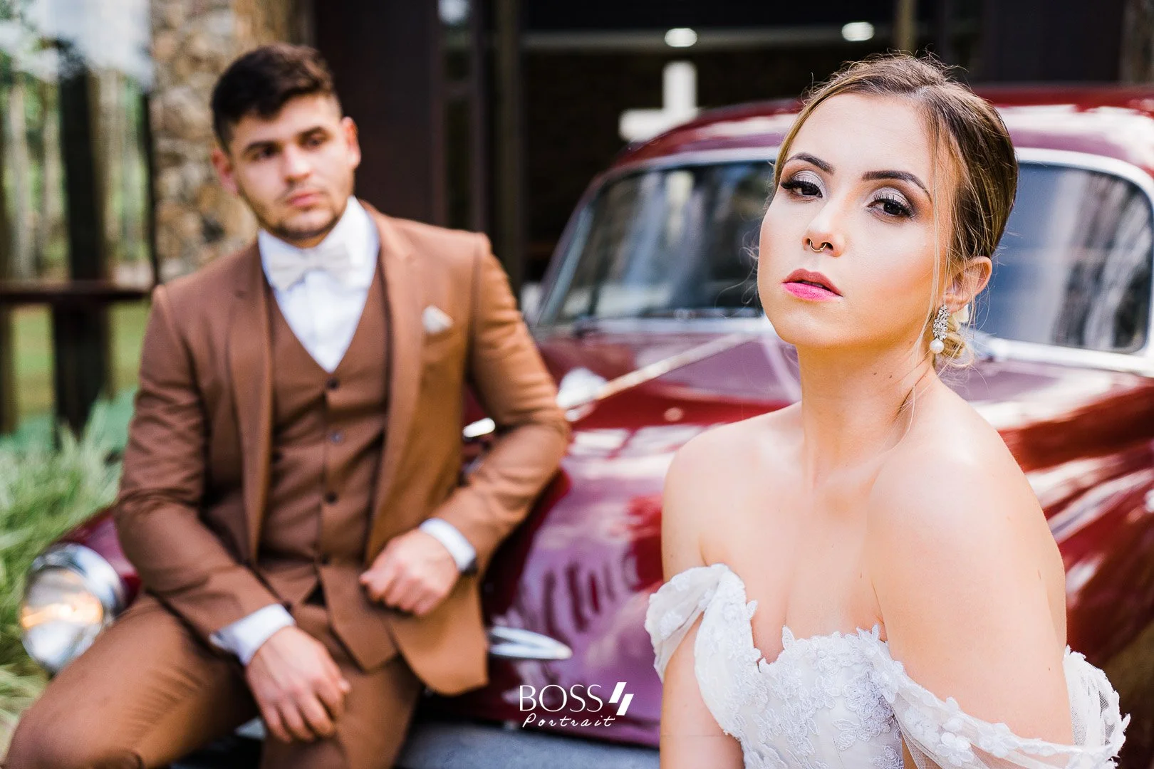 Casal de noivos em traje elegante posando ao lado de um carro antigo vermelho, ao ar livre.