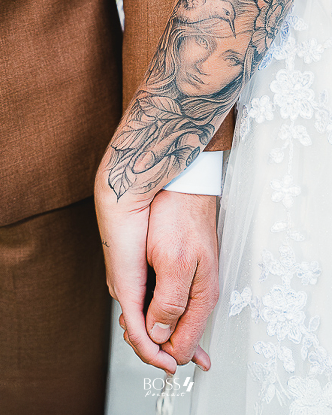 Mãos entrelaçadas de um casal, com uma tatuagem visível no braço de uma das pessoas, vestindo roupa de casamento.