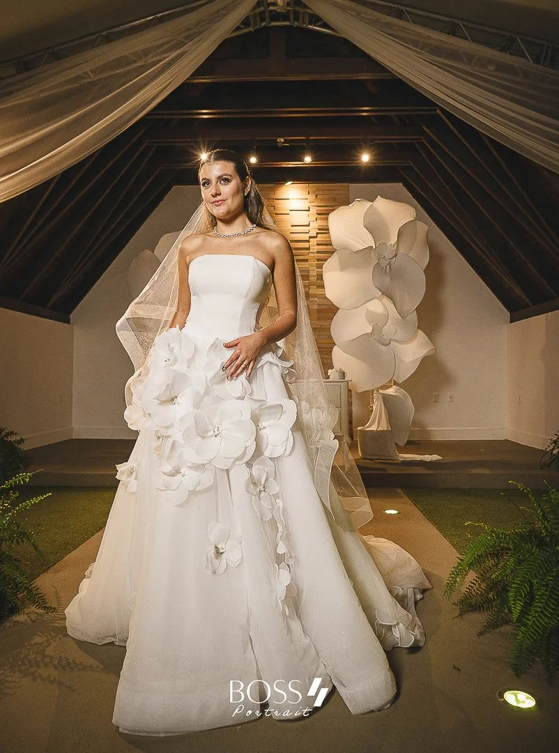 Noiva de vestido branco com detalhes de flores, usando véu e joias, posando dentro de uma estrutura de madeira decorada com plantas verdes e grandes flores brancas.