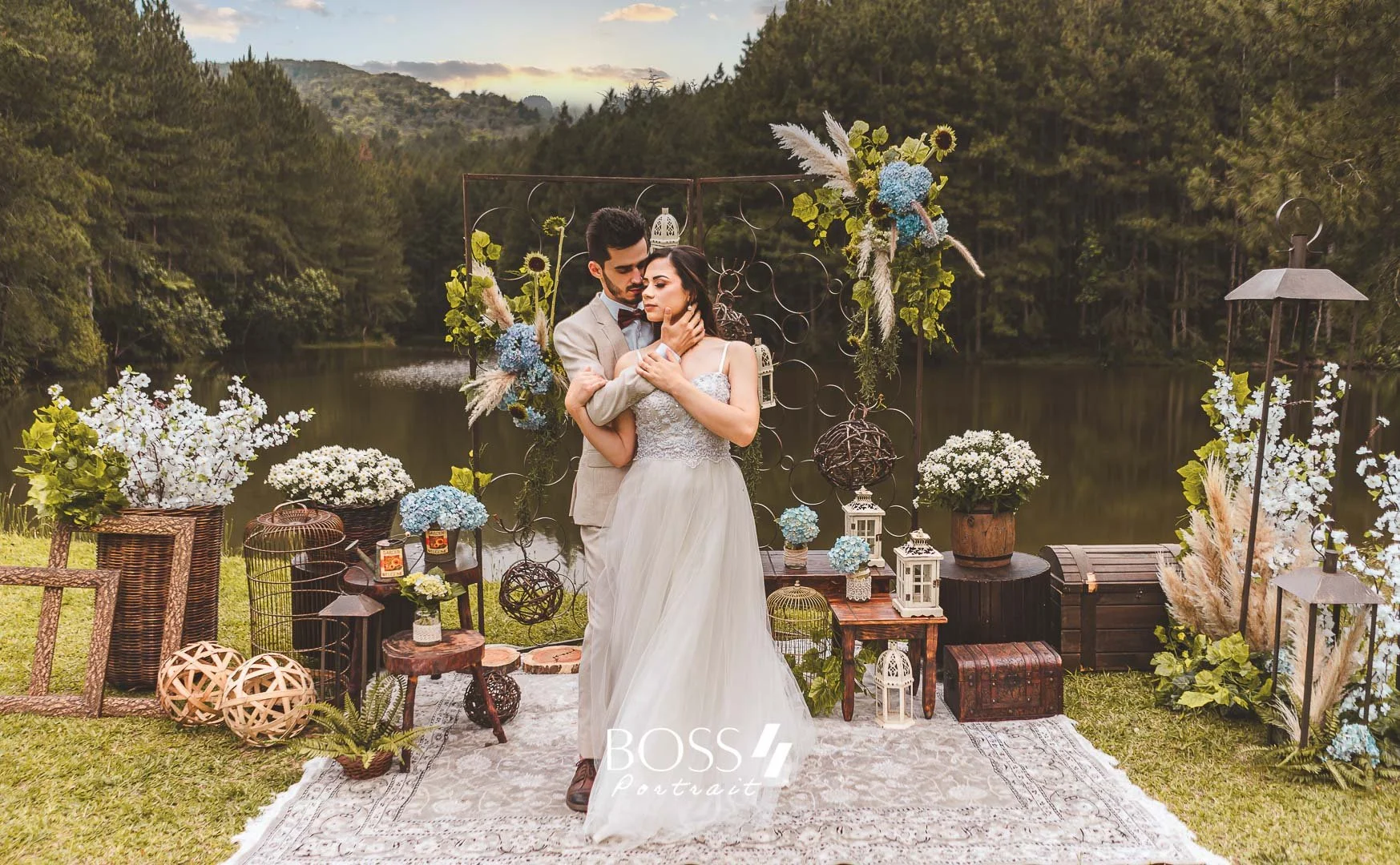 Casal de casamento em festa ao ar livre, com decoração de flores e objetos rústicos, próximo a um lago em ambiente natural, ao entardecer.