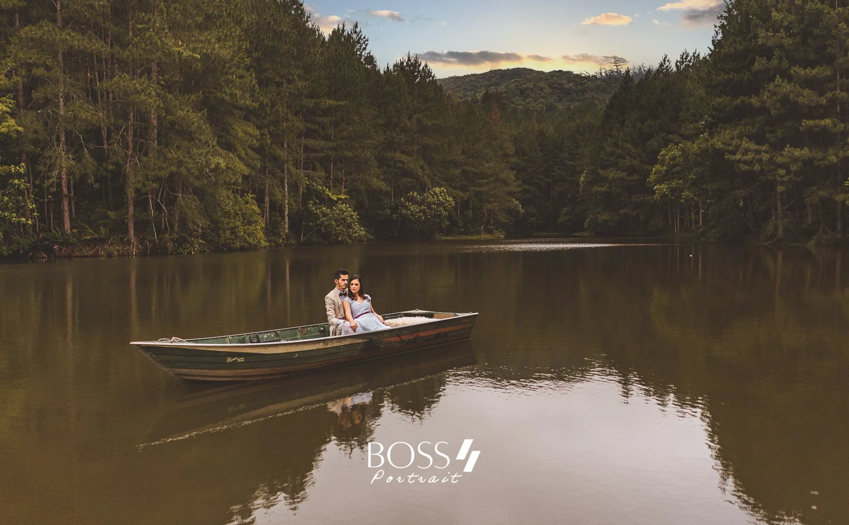 Casal sentado em um barco em um rio cercado por uma floresta densa. O céu ao entardecer apresenta nuvens e uma tonalidade suave, refletida na água tranquila do rio.