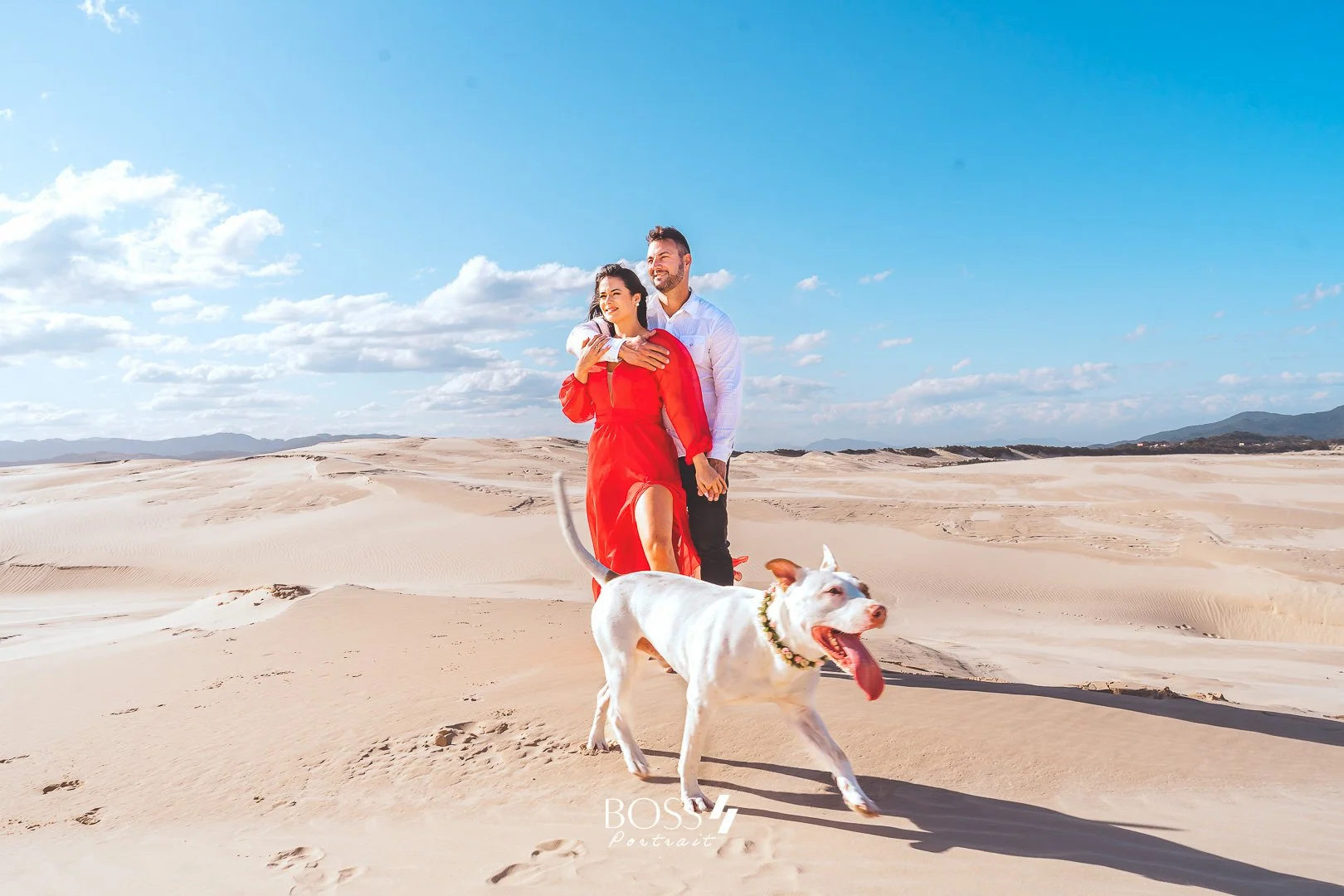 Casal caminhando com cachorro em deserto de dunas sob céu azul com nuvens.