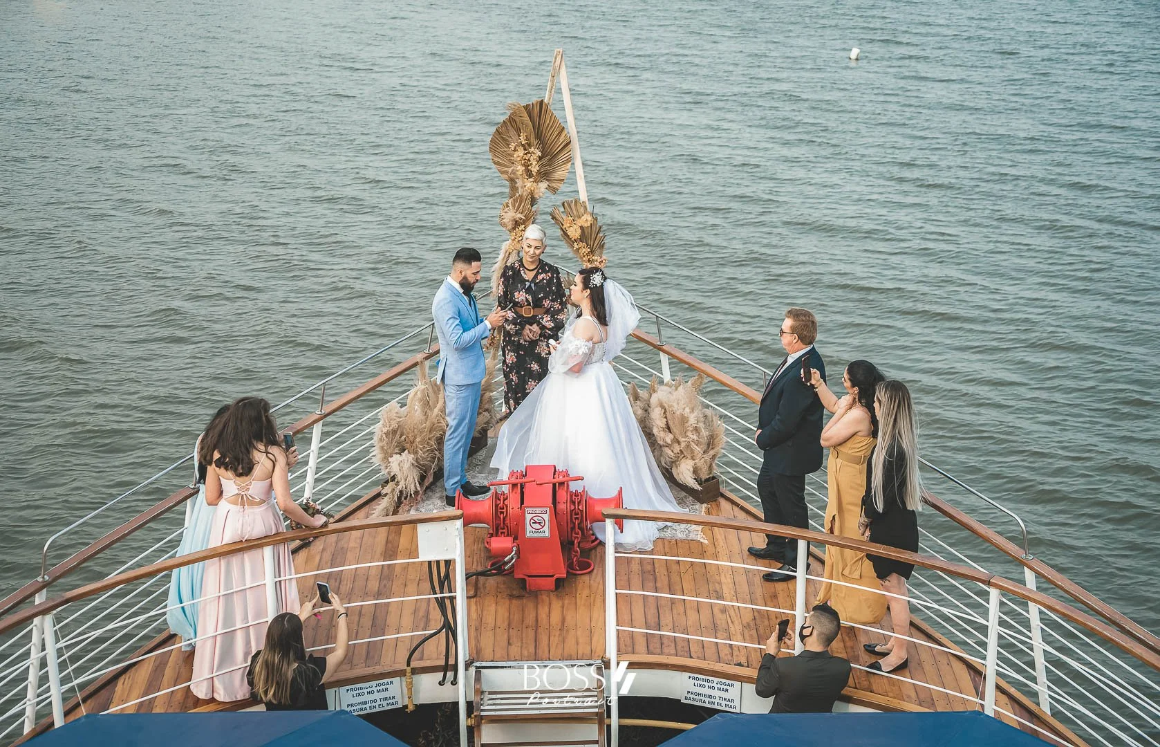 Casamento a bordo de um barco com o casal principal vestido de branco, rodeado por convidados na embarcação, com o mar ao fundo.