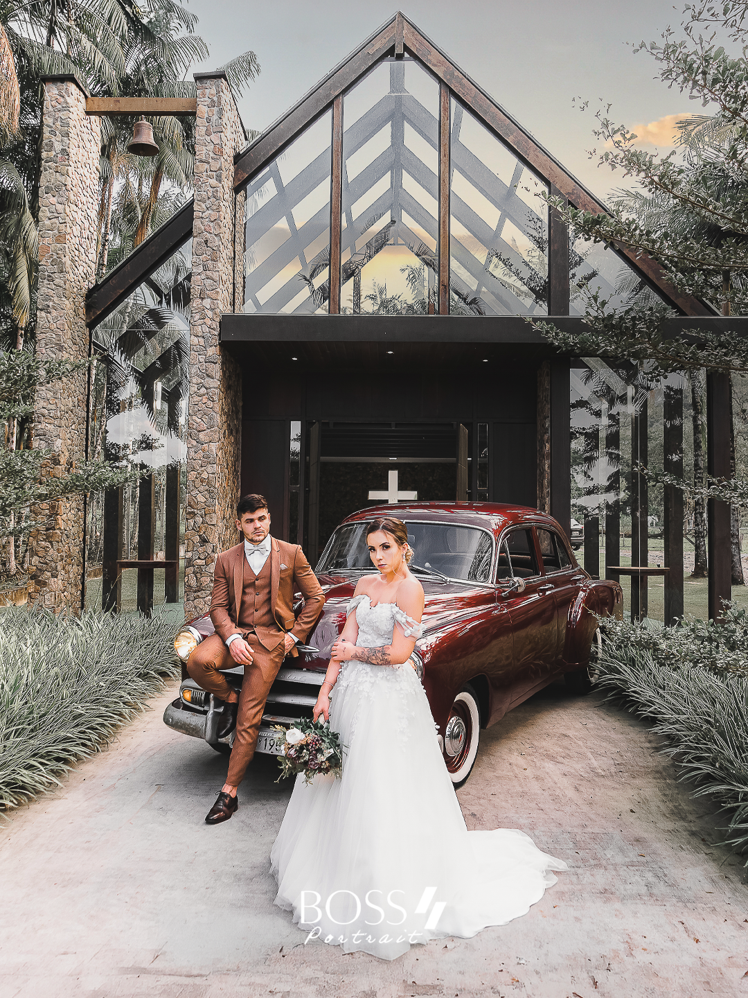 Casal de noivos posando na frente de um carro antigo, com uma capela de fundo, rodeados de árvores e plantas, durante o pôr do sol.