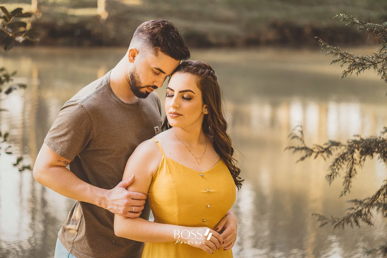 Um casal está junto perto de um rio ao pôr do sol, com árvores ao redor, e a mulher veste um vestido amarelo enquanto o homem usa uma camiseta cinza.