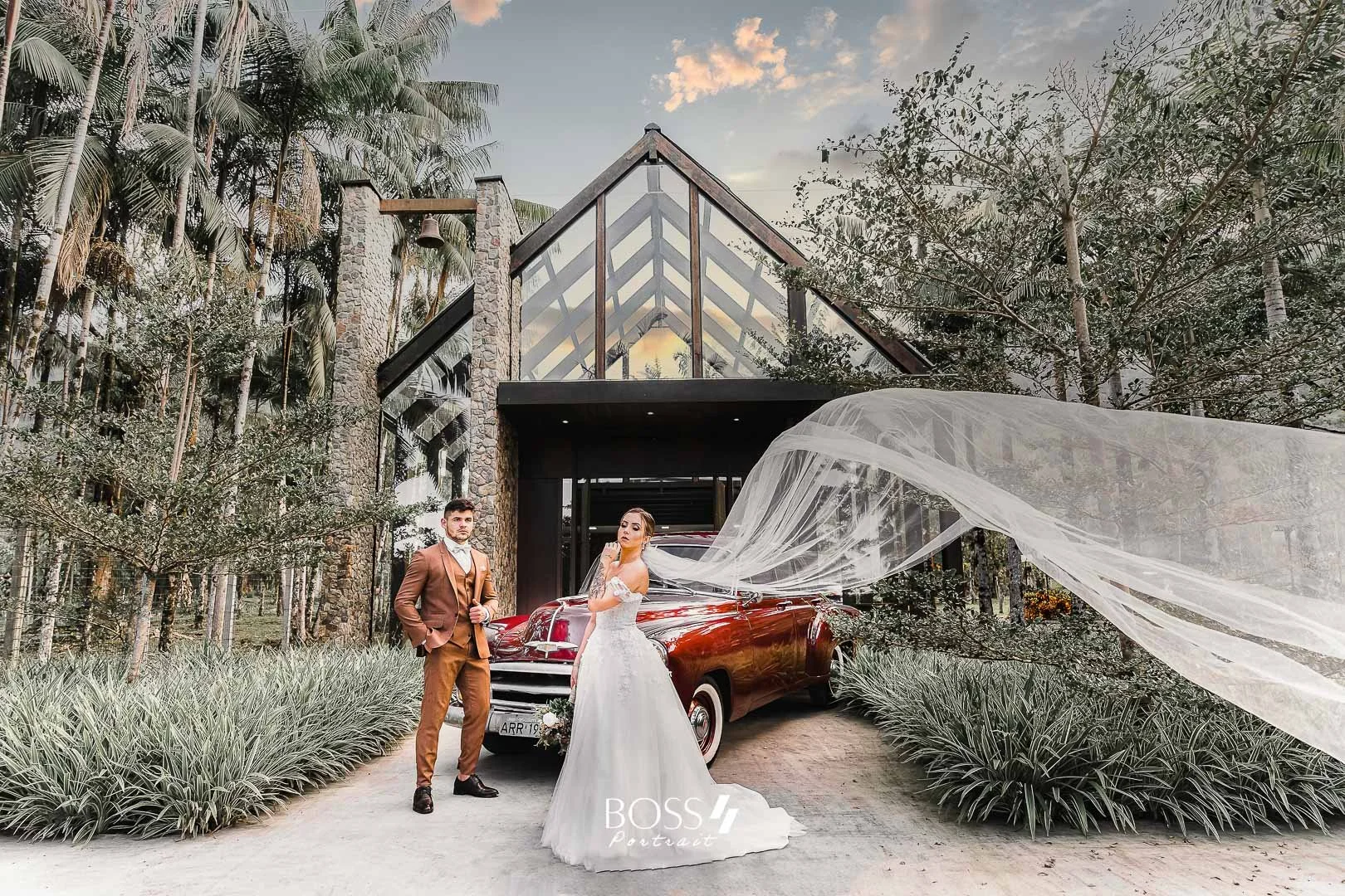 Casal de noivos posando na frente de um carro clássico vermelho, ao lado de uma casa moderna de estrutura de vidro e pedra, cercada por árvores e plantas, em uma ocasião de casamento ao ar livre.