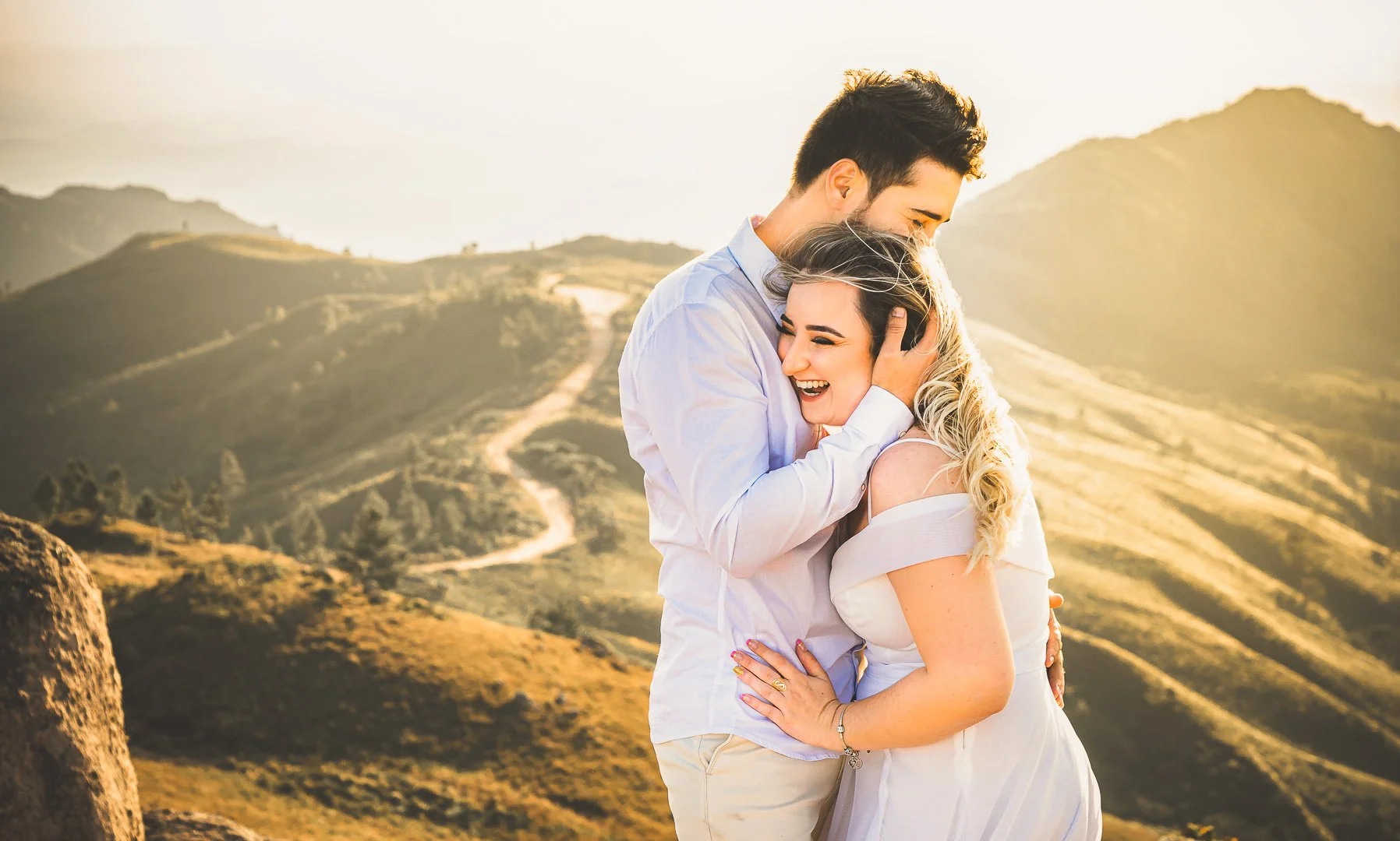 Um casal sorridente abraçado em um pôr do sol, em uma paisagem de colinas e caminho de terra