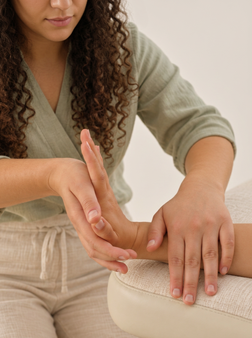 A massage therapist performs a massage on a person's arm and hand, sitting on a beige cushioned surface.