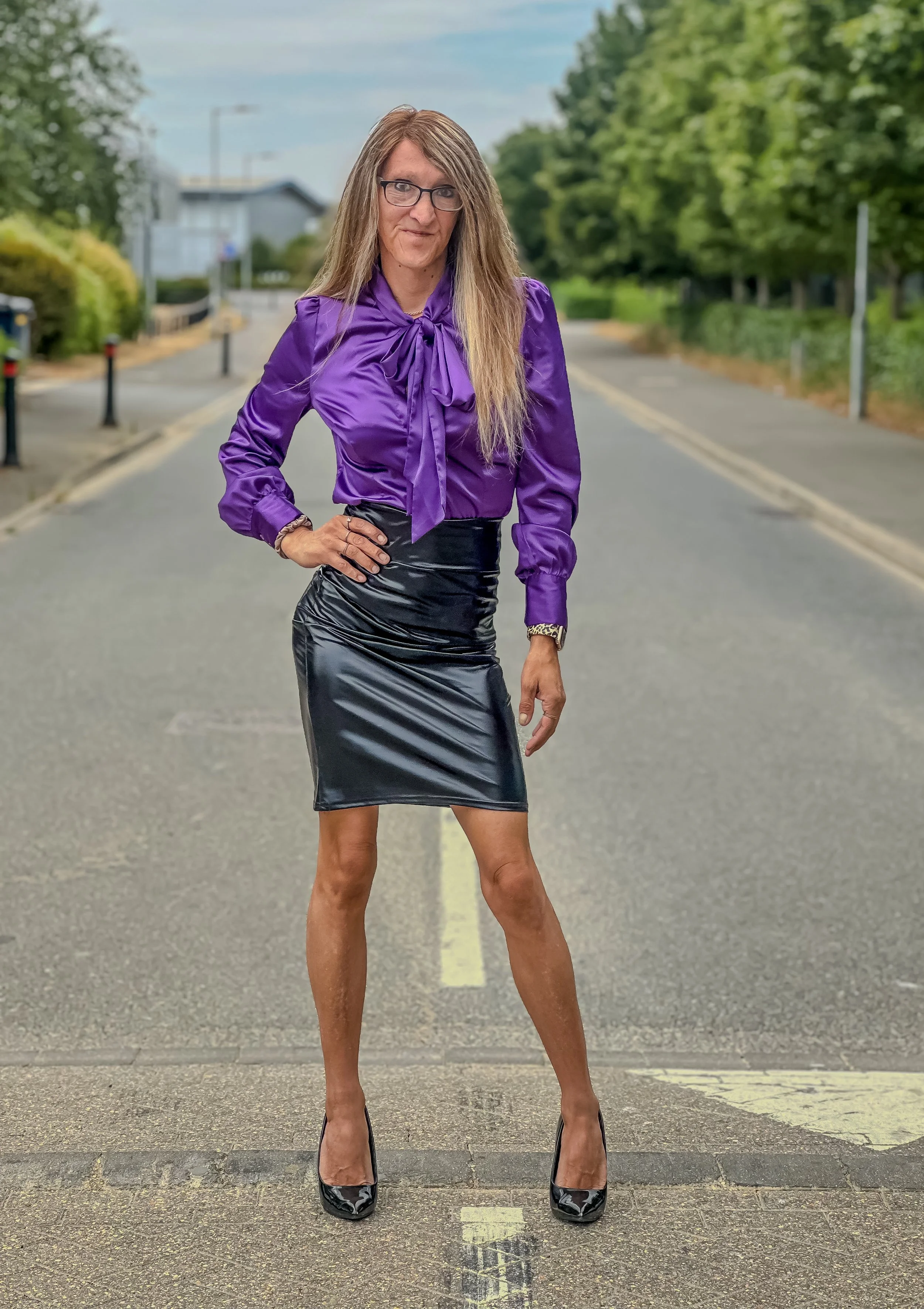 A woman standing in the middle of a road with trees and houses in the background, wearing a purple blouse, a black leather skirt, and high heels.