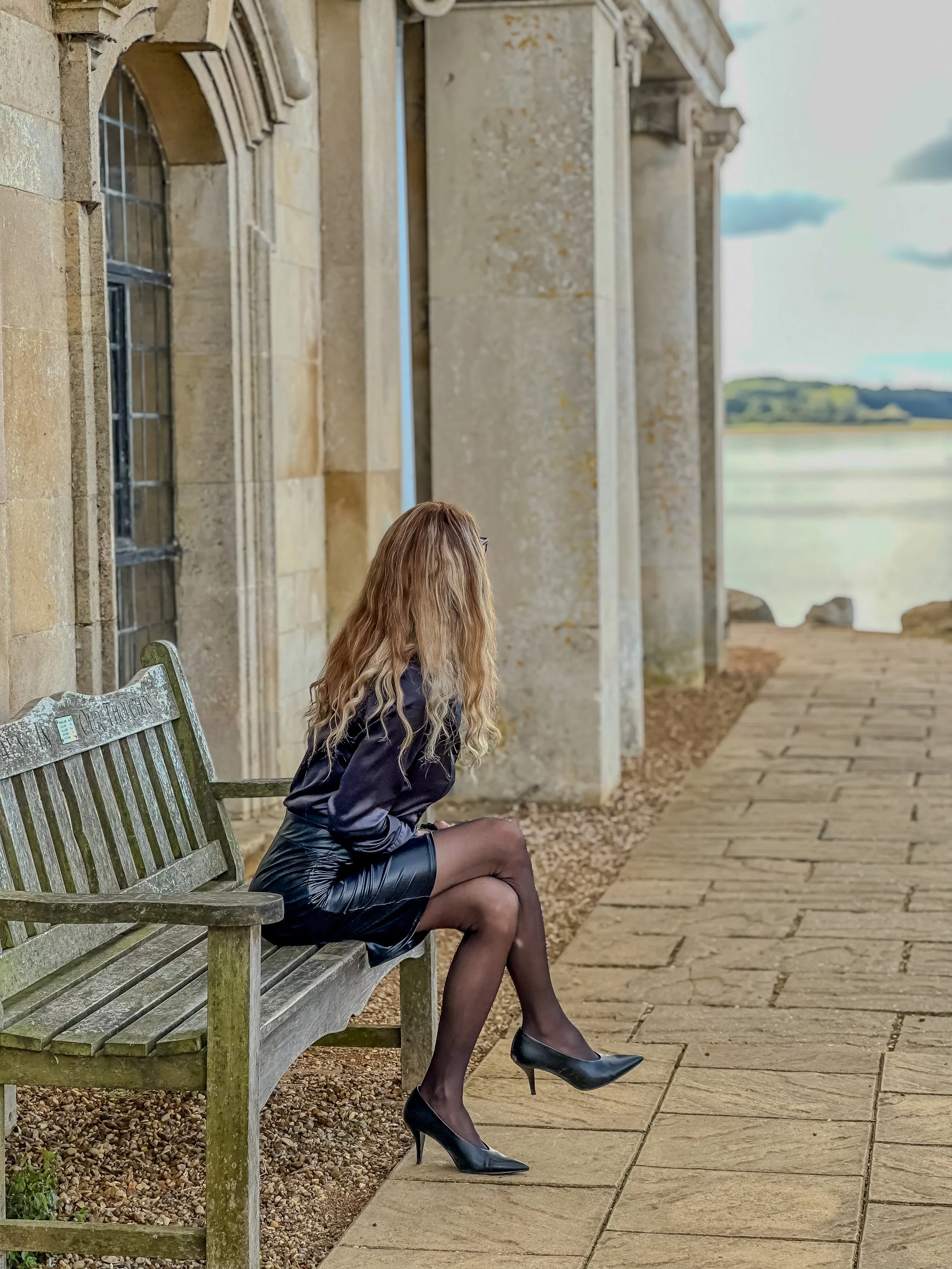 A woman with long, wavy hair sitting on a wooden bench outside beside a stone building with arched windows and tall columns. She is wearing a black dress, black sheer stockings, and black high heels. The background shows an outdoor pathway and distan