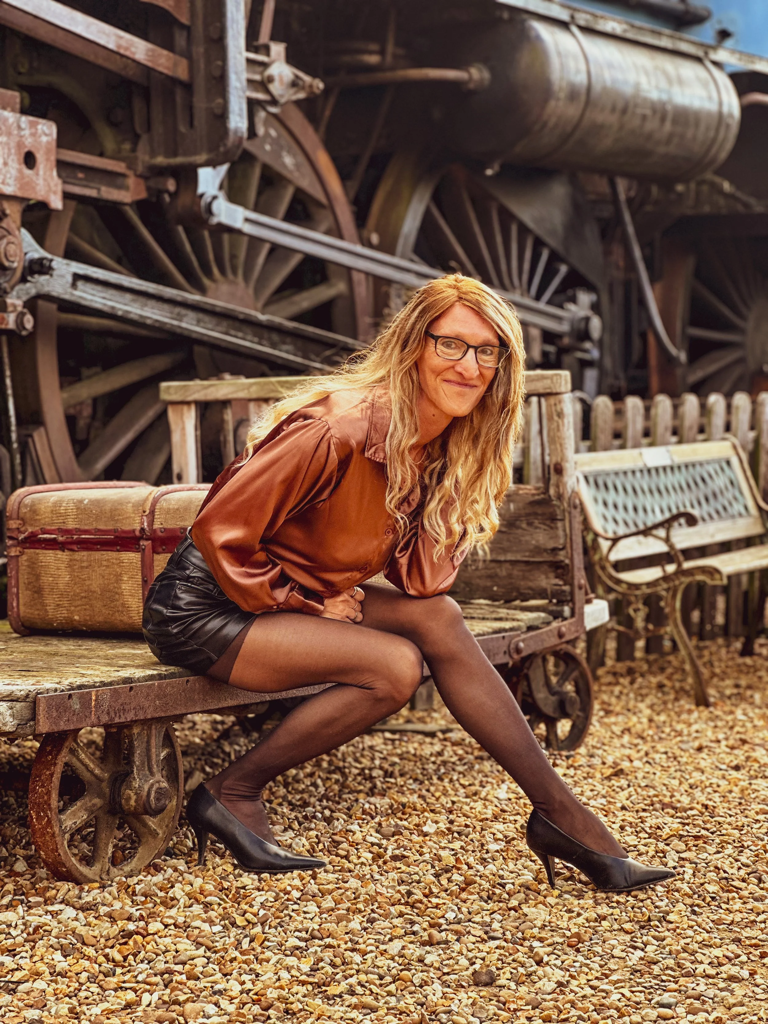 A woman with long blonde hair, glasses, wearing a brown satin shirt, black leather shorts, thigh-high stockings, and high heels, sitting on a wooden bench outdoors with a vintage train engine in the background.