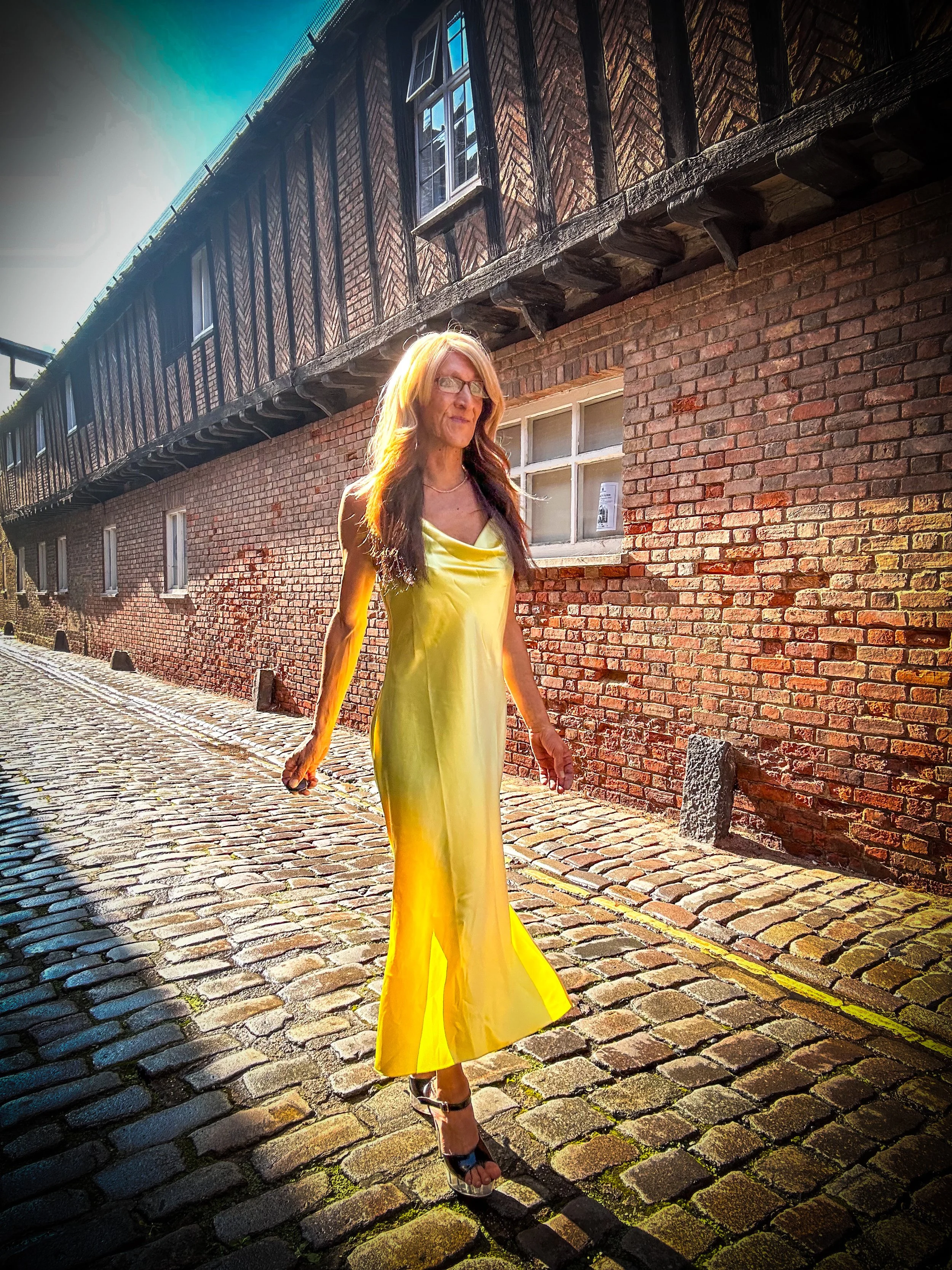 A woman with long hair and glasses walking on a cobblestone street in front of a brick building with windows. She is wearing a long, yellow satin dress and high heels, and the sunlight is highlighting her hair.