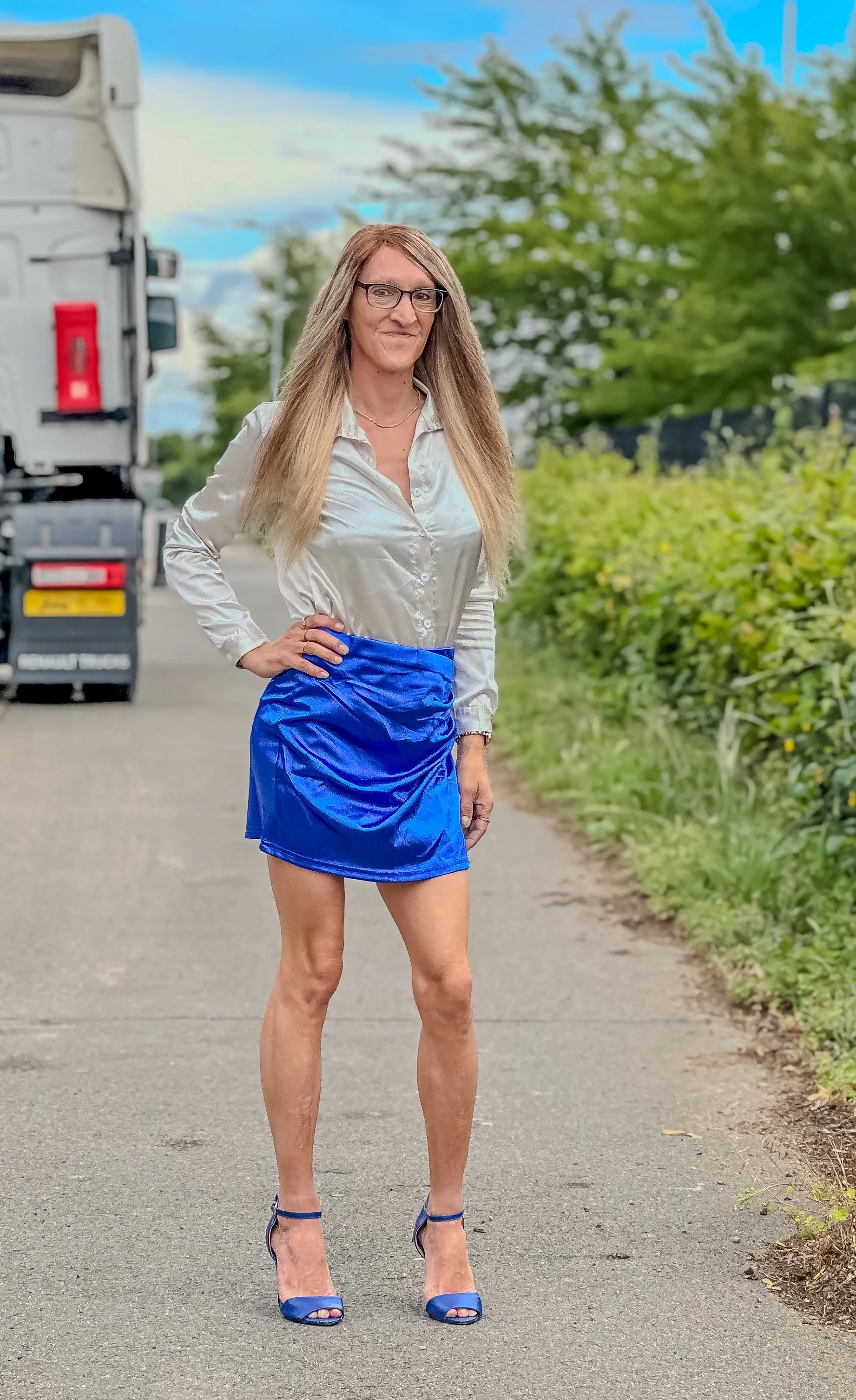 A woman with long blonde hair, wearing glasses, a shiny white blouse, a bright blue skirt, and matching blue high heels, standing outdoors on a paved path with greenery and a truck in the background.