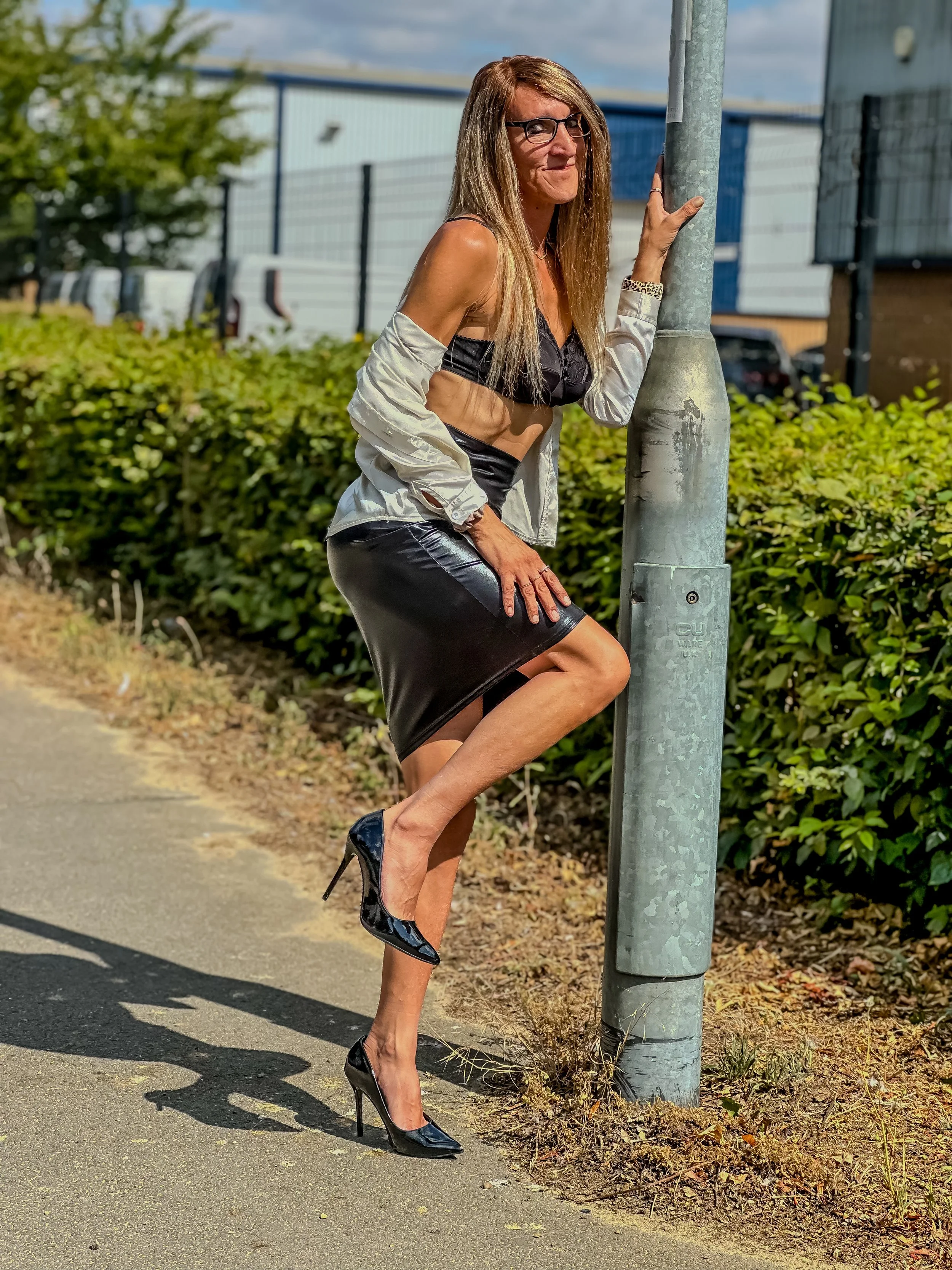 A woman with long brown hair, glasses, wearing a black bra, shiny black skirt, a shiny jacket hanging off her shoulders, and high heels, posing outdoors while holding onto a metal pole.