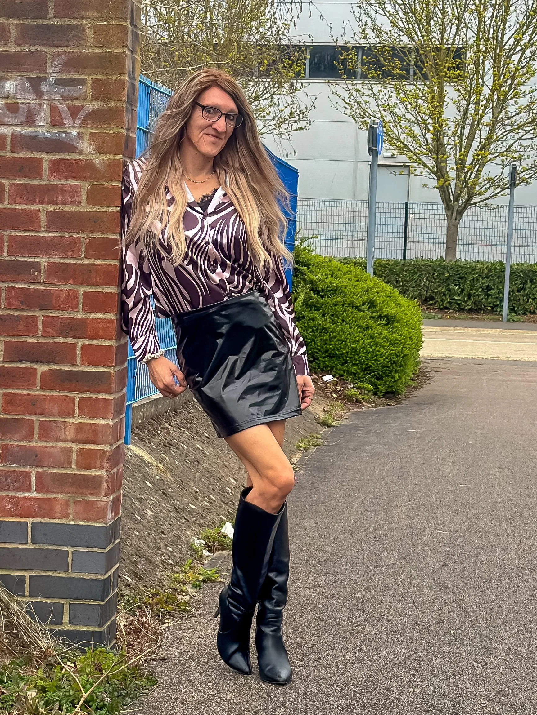 A woman with long blonde hair, glasses, and a patterned shirt leaning against a brick wall outdoors. She is wearing a black shiny skirt and tall black boots, standing on a paved sidewalk next to bushes, with trees and a white vehicle in the backgroun