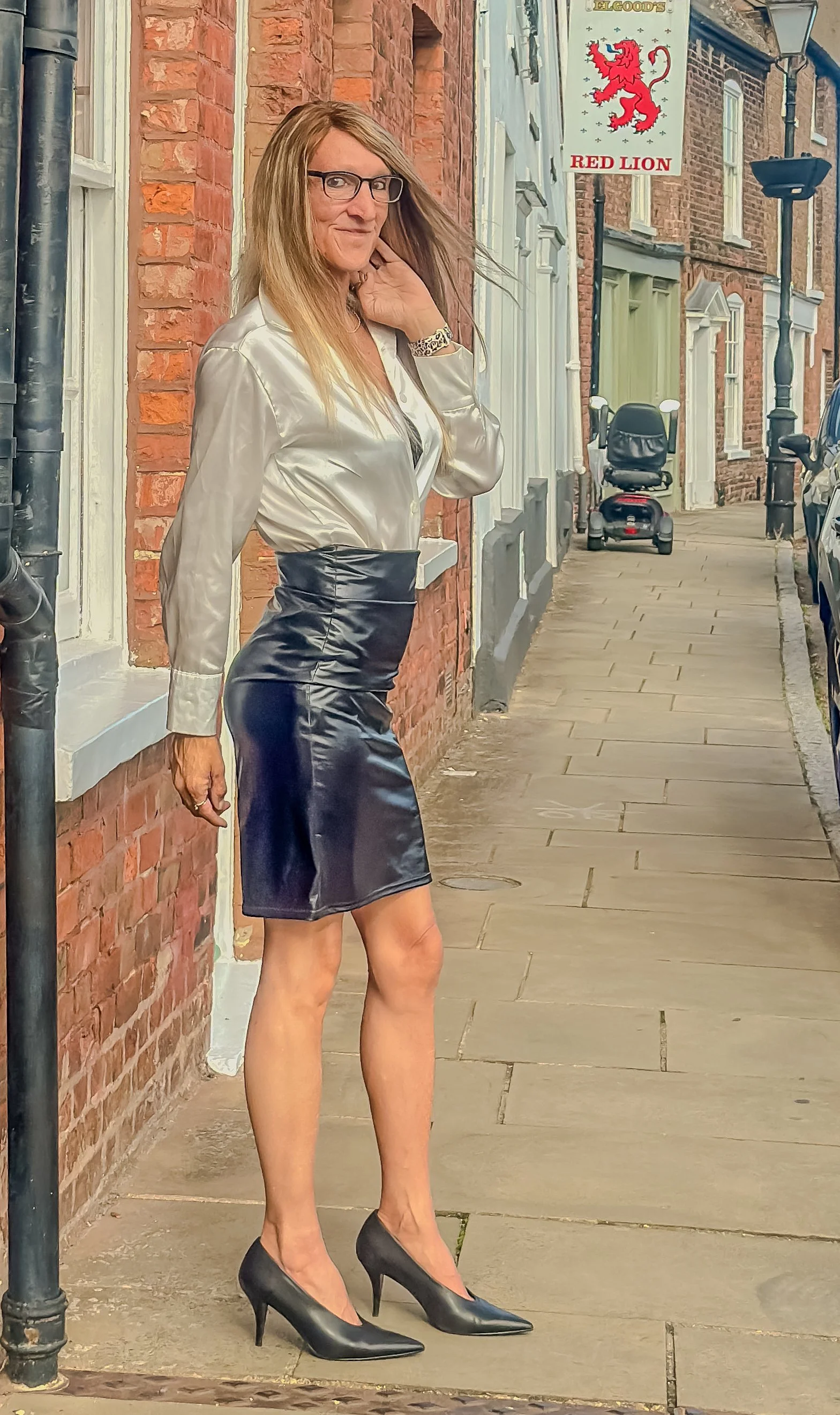 A woman with long blonde hair, glasses, and a satin blouse posing on a sidewalk next to brick buildings wearing a leather skirt and black high heels.