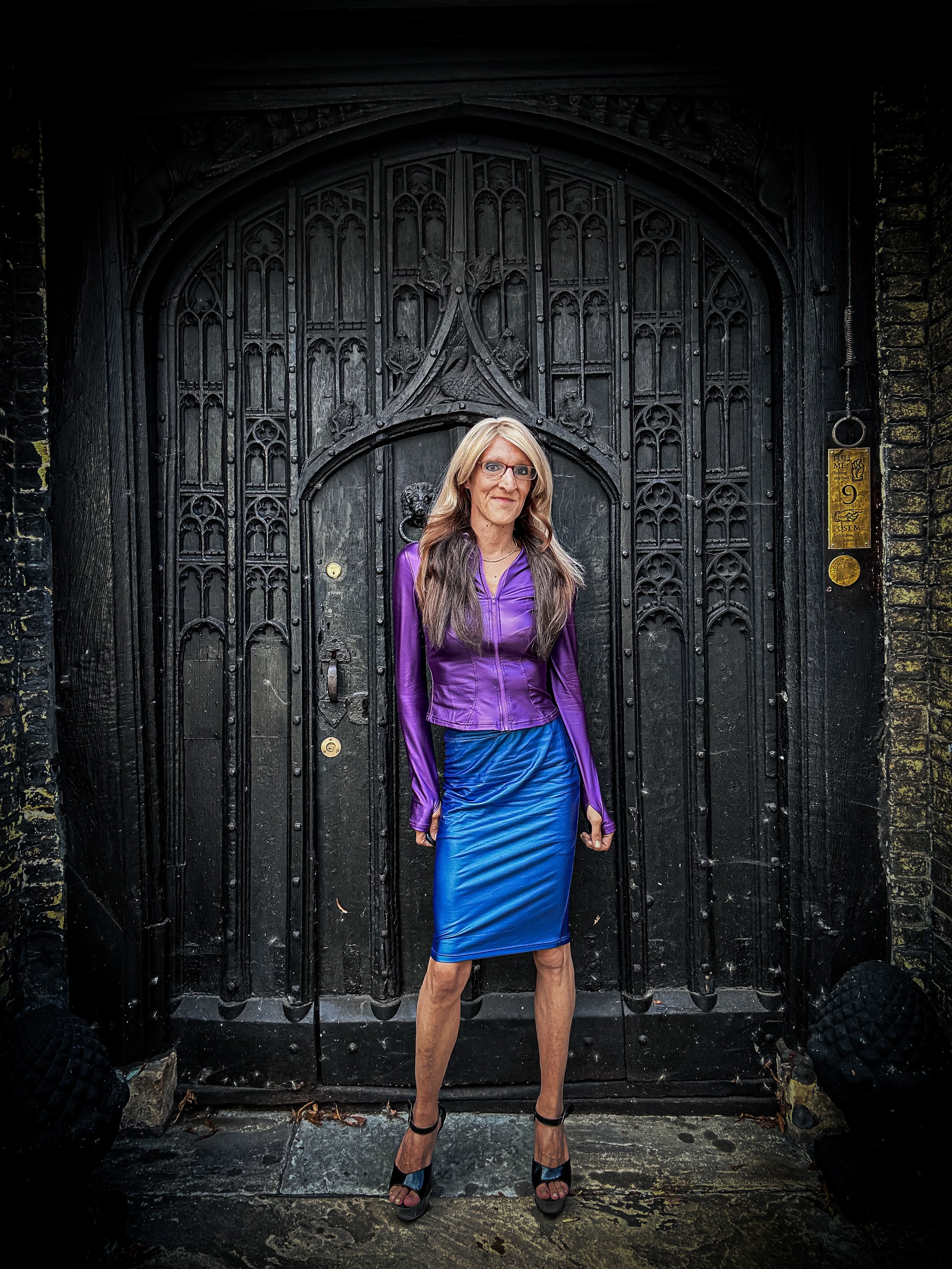 A woman with long blond hair wearing a purple jacket, blue skirt, and black high heels standing in front of a decorative black wooden door with ornate carvings.