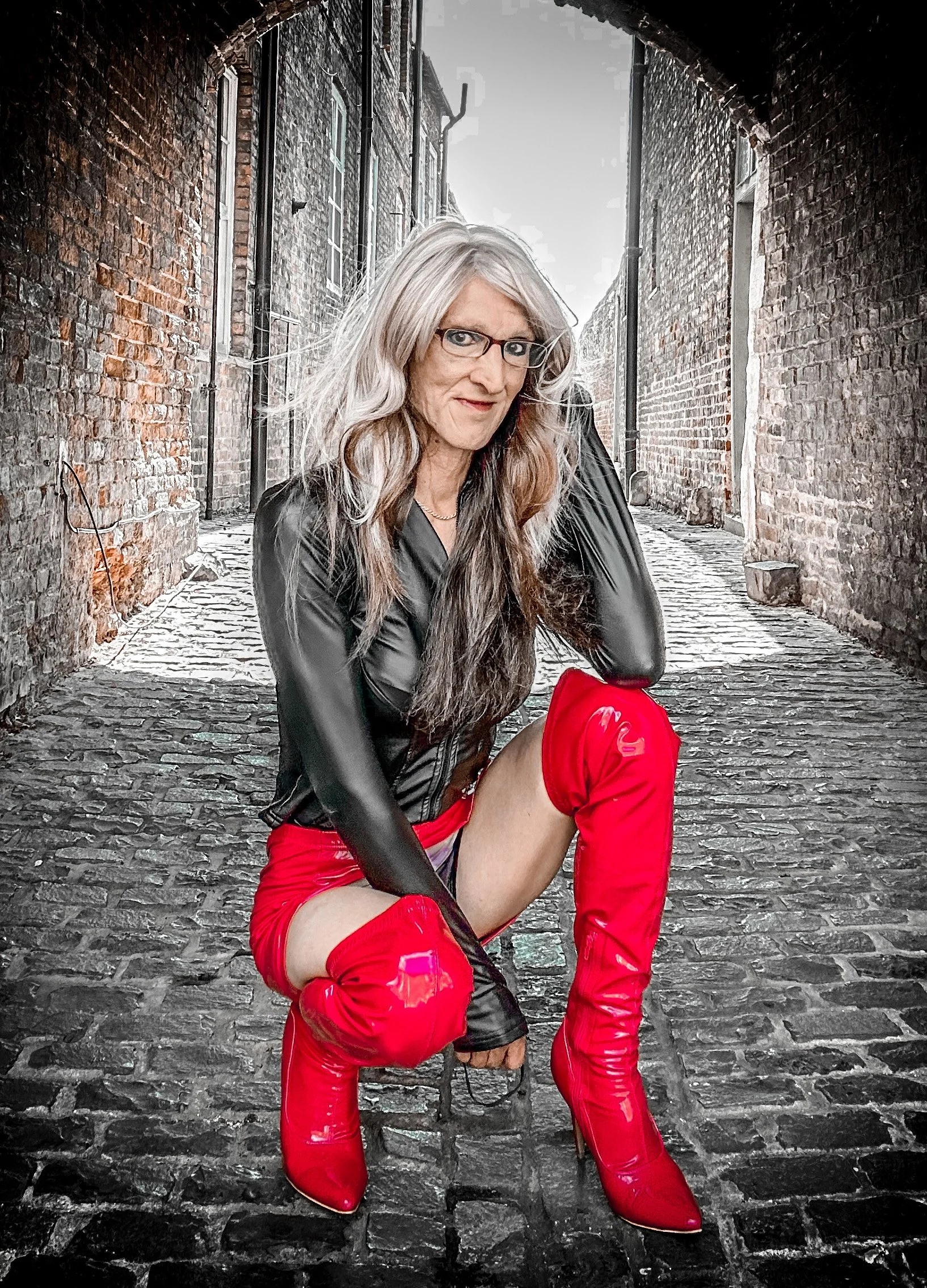 A woman with long gray hair, glasses, and a black leather jacket, wearing red thigh-high boots, sitting on cobblestone street between brick walls in an alleyway.