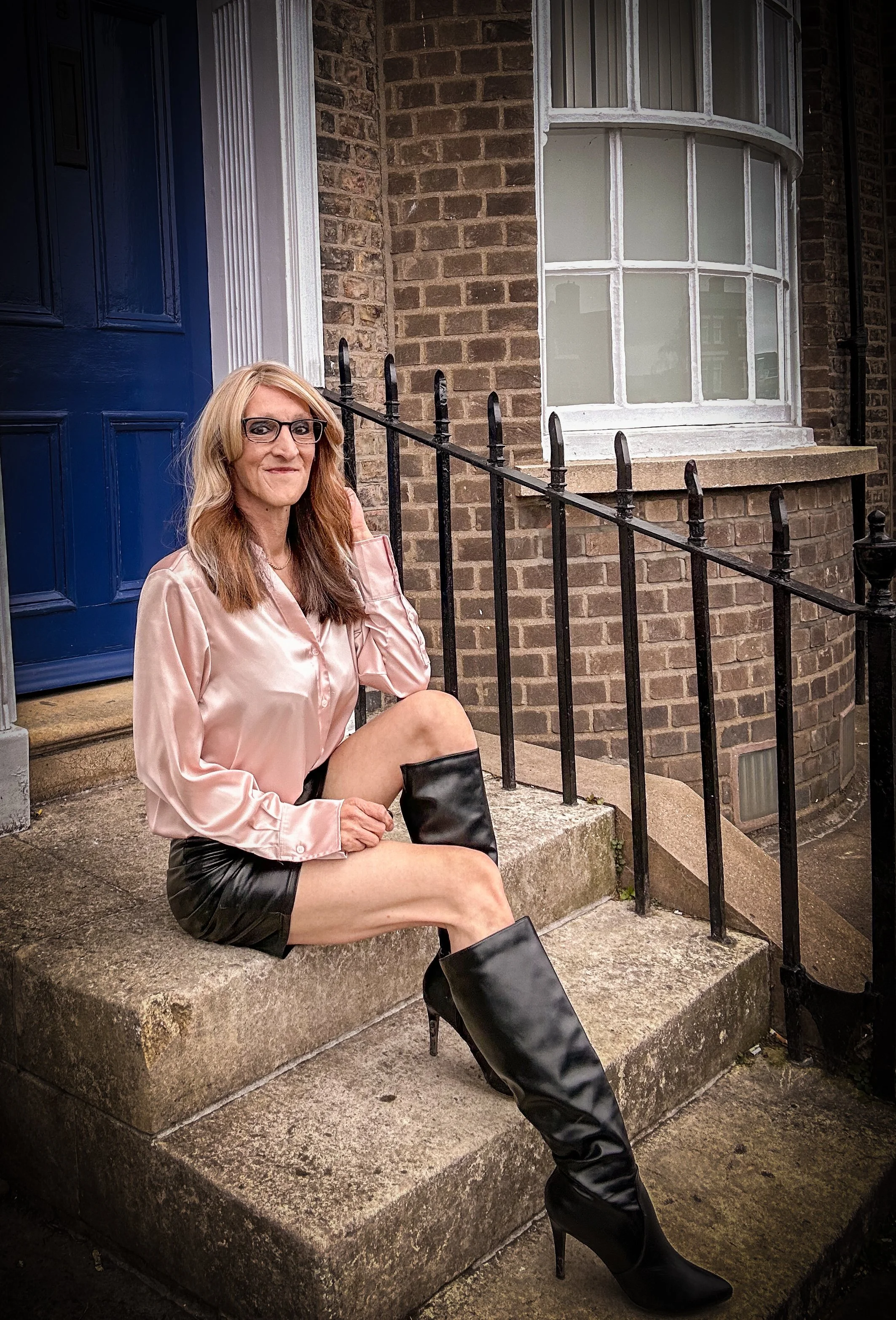 A woman sitting on the front steps of a brick house with a blue door and a large bay window. She has shoulder-length blonde hair, glasses, and is wearing a shiny pink blouse, black leather shorts, and black high-heeled boots.