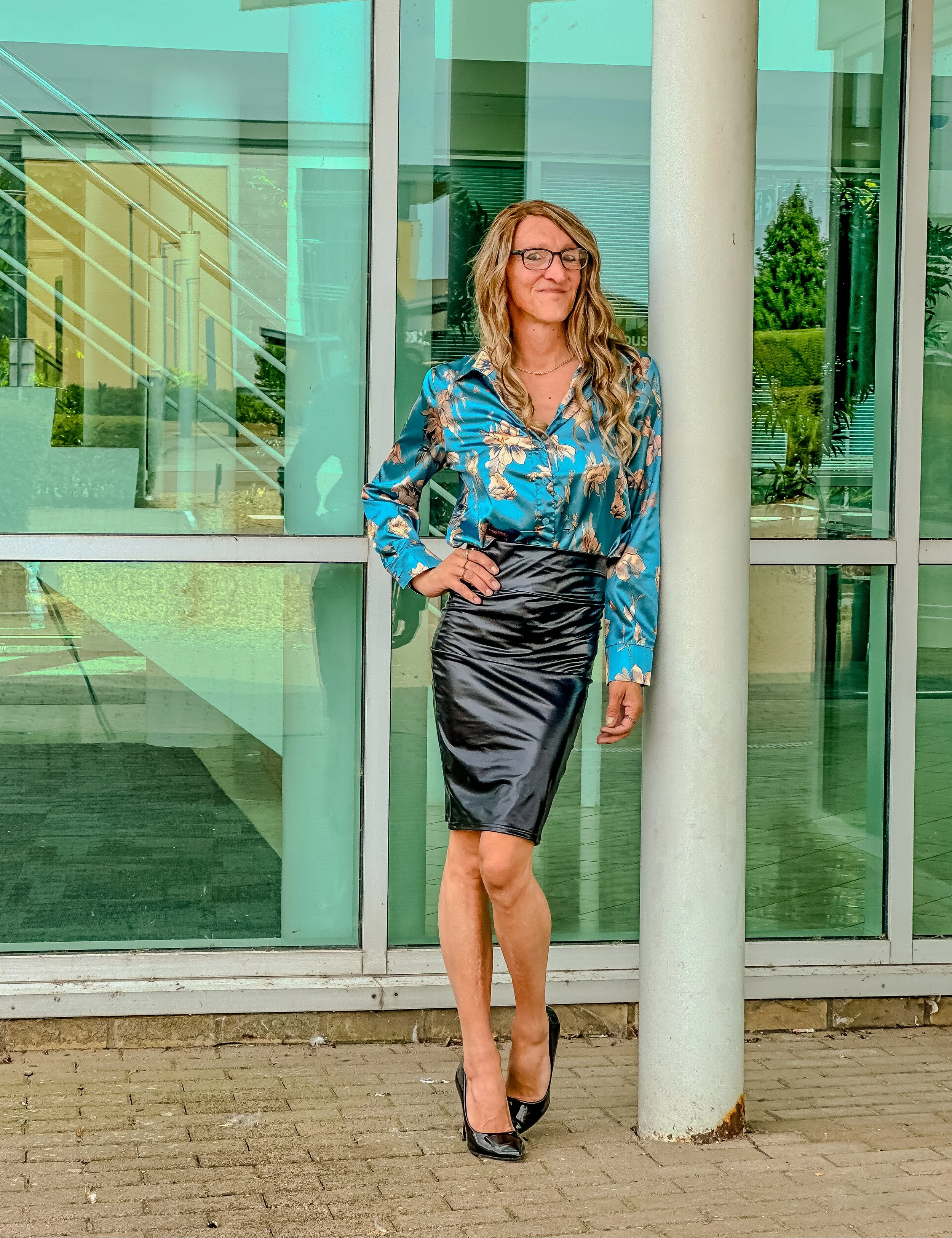 A woman with long wavy blonde hair and glasses is standing outdoors in front of a glass building. She is wearing a blue floral blouse, a black shiny leather skirt, and black high heels. She is posing with one hand on her hip and smiling slightly.
