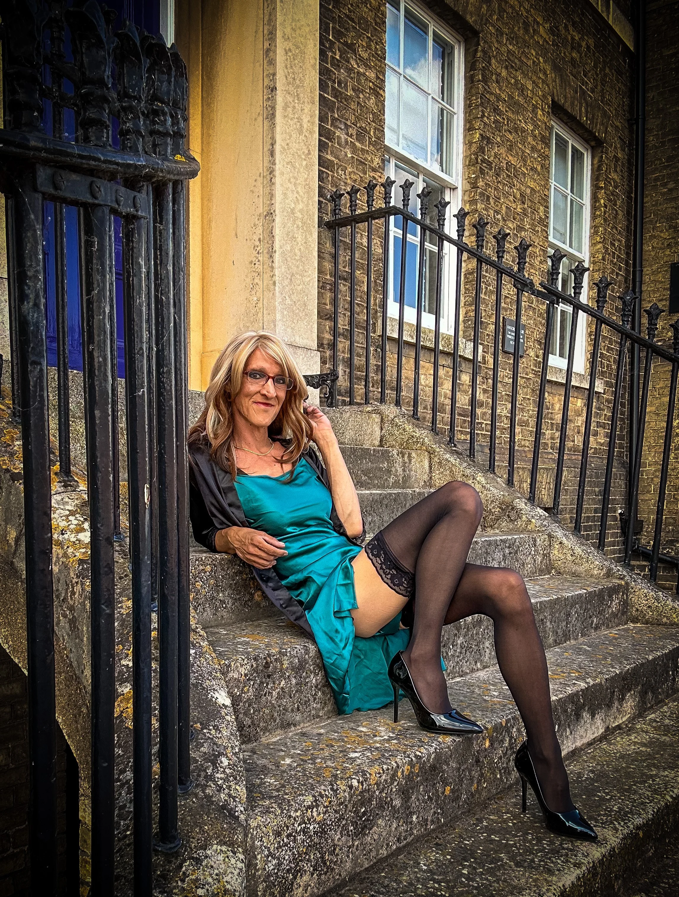 A woman with blonde hair, glasses, wearing black high heels, black stockings, and a teal dress, sitting on outdoor stone steps beside a black metal railing, with a brick building in the background.